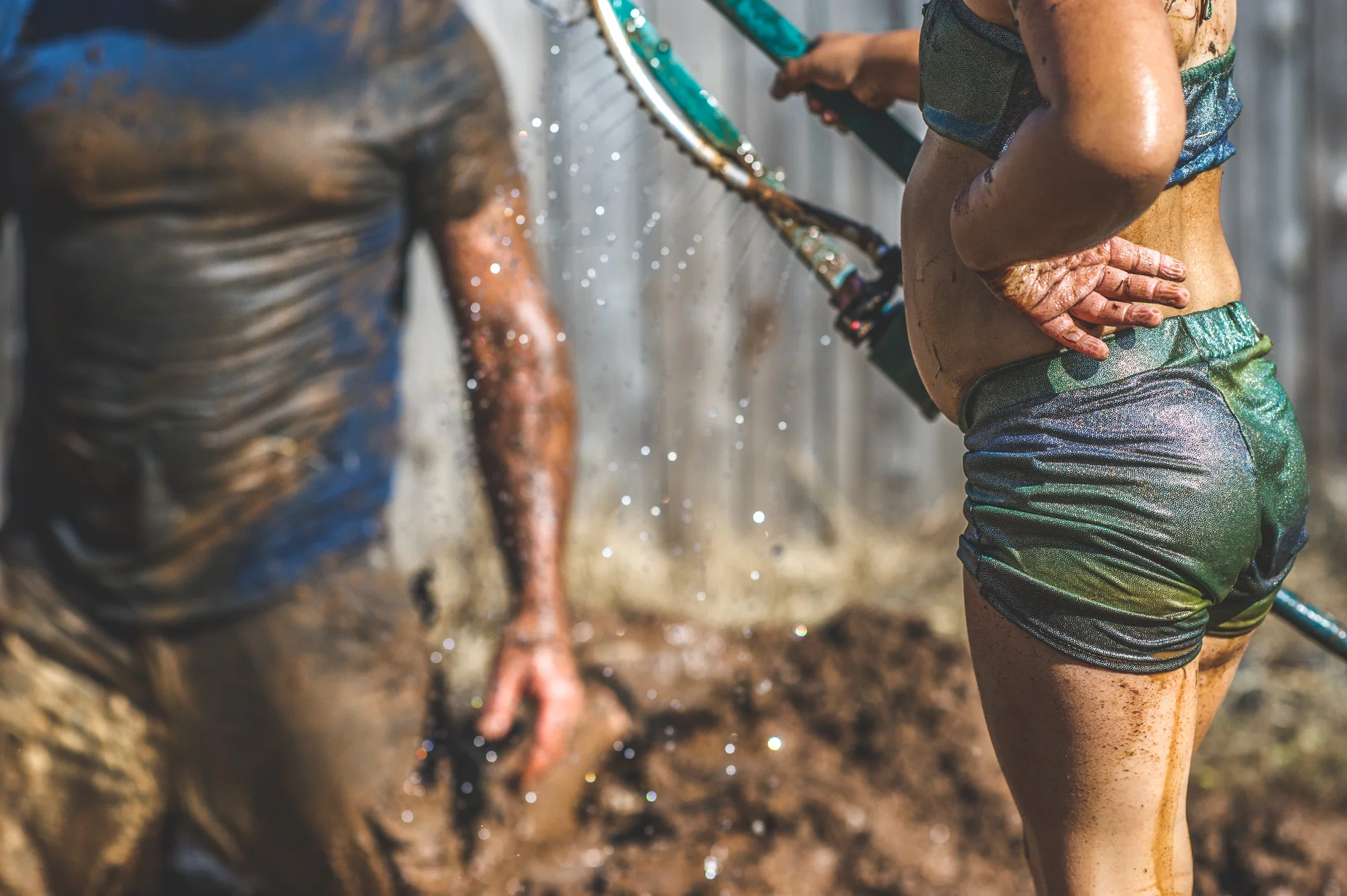 Color photo of a little girl in a green, sparkly bathing suit putting water from the sprinkler into a mud hole as her father watches on