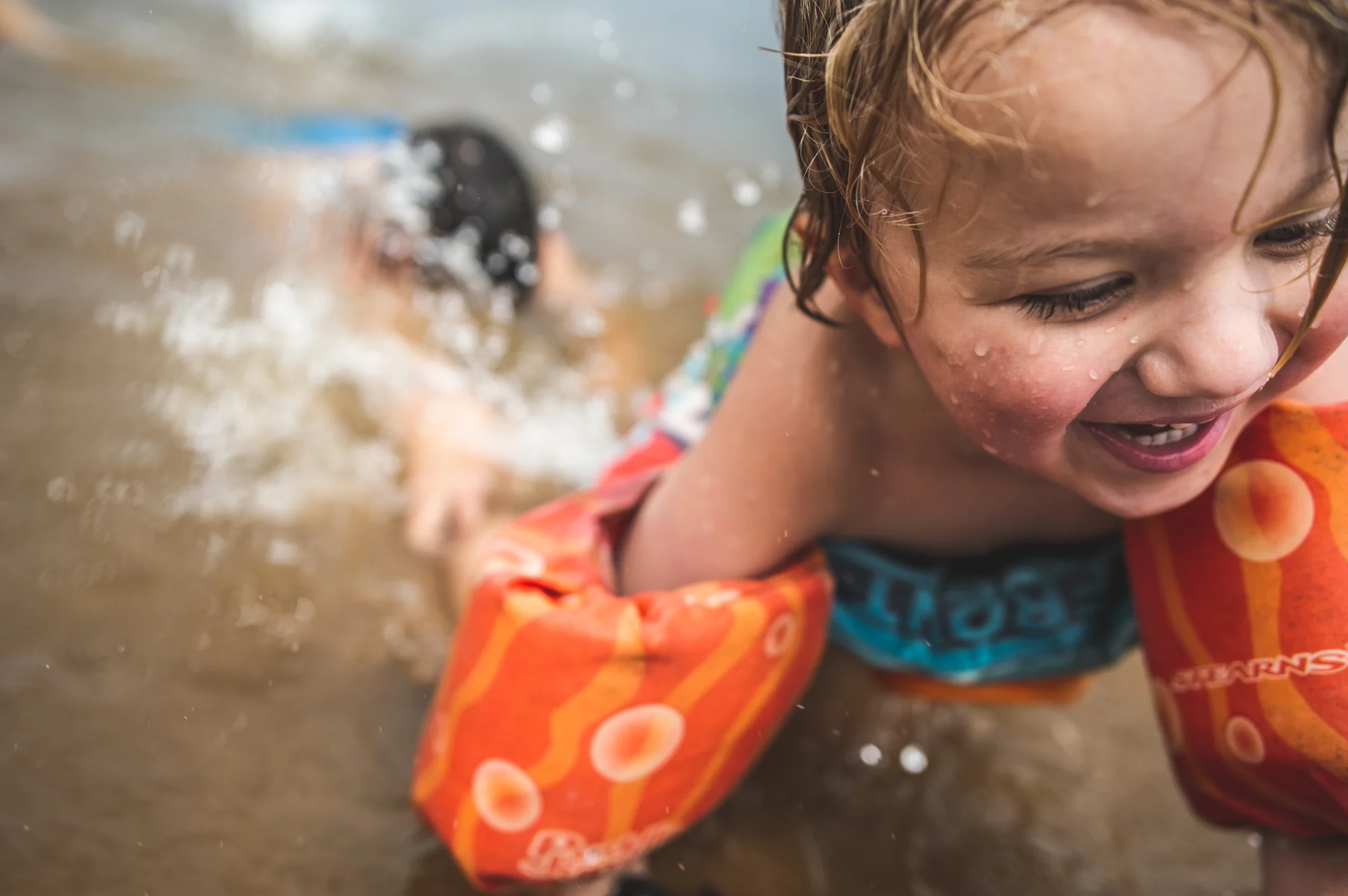 Color photo of a young boy with floaties on climbing out of a lake while his brother grabs his legs and tries to pull him back in