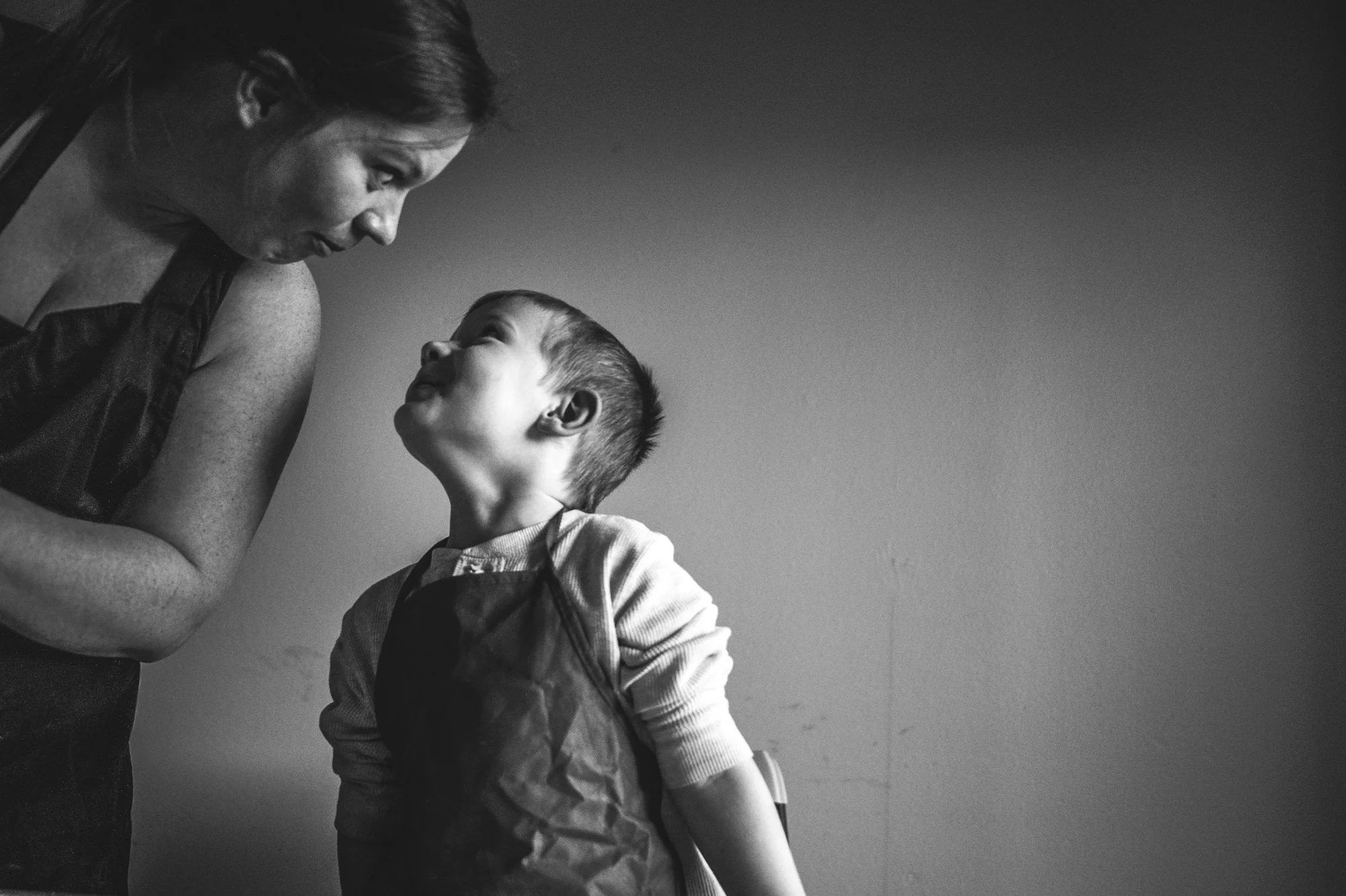 Black and white photo of a mother and son, both wearing aprons, and staring at each other, giving each other goofy looks in Aurora, CO
