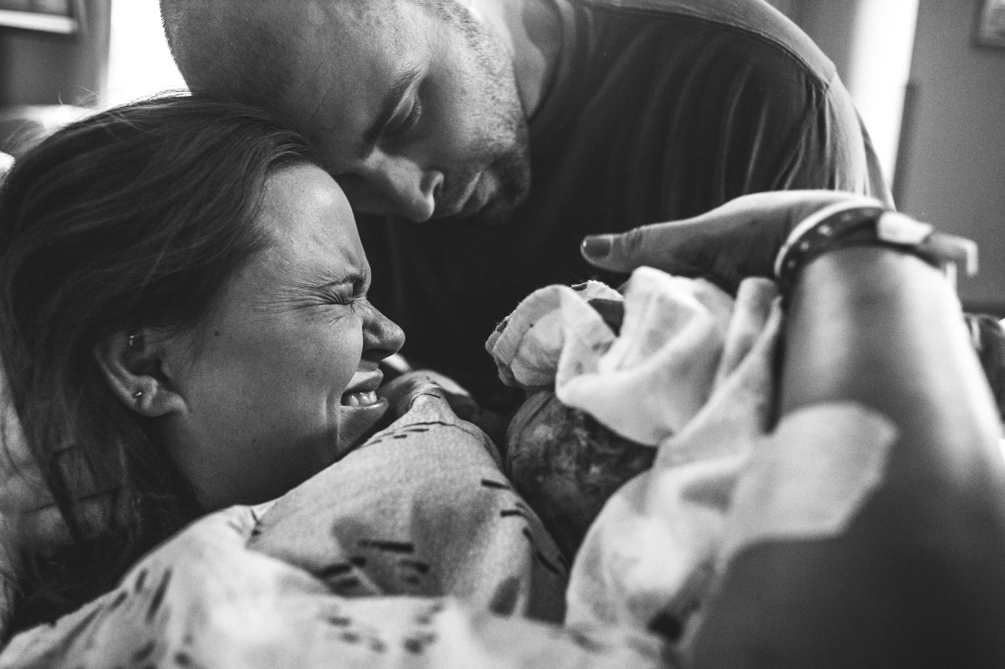 Black and white photo of a mother and father meeting their newborn daughter for the first time in the hospital. The father lightly places his forehead on his wife's while his wife holds back tears of joy