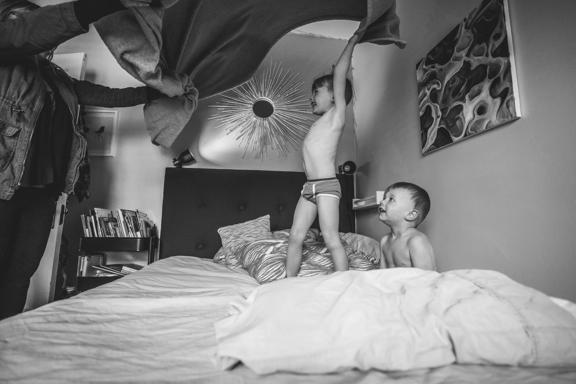 Black and white photo of a little boy and his brother on their bed as their mother waves a blanket over their heads in their home in Park Hill, Colorado.