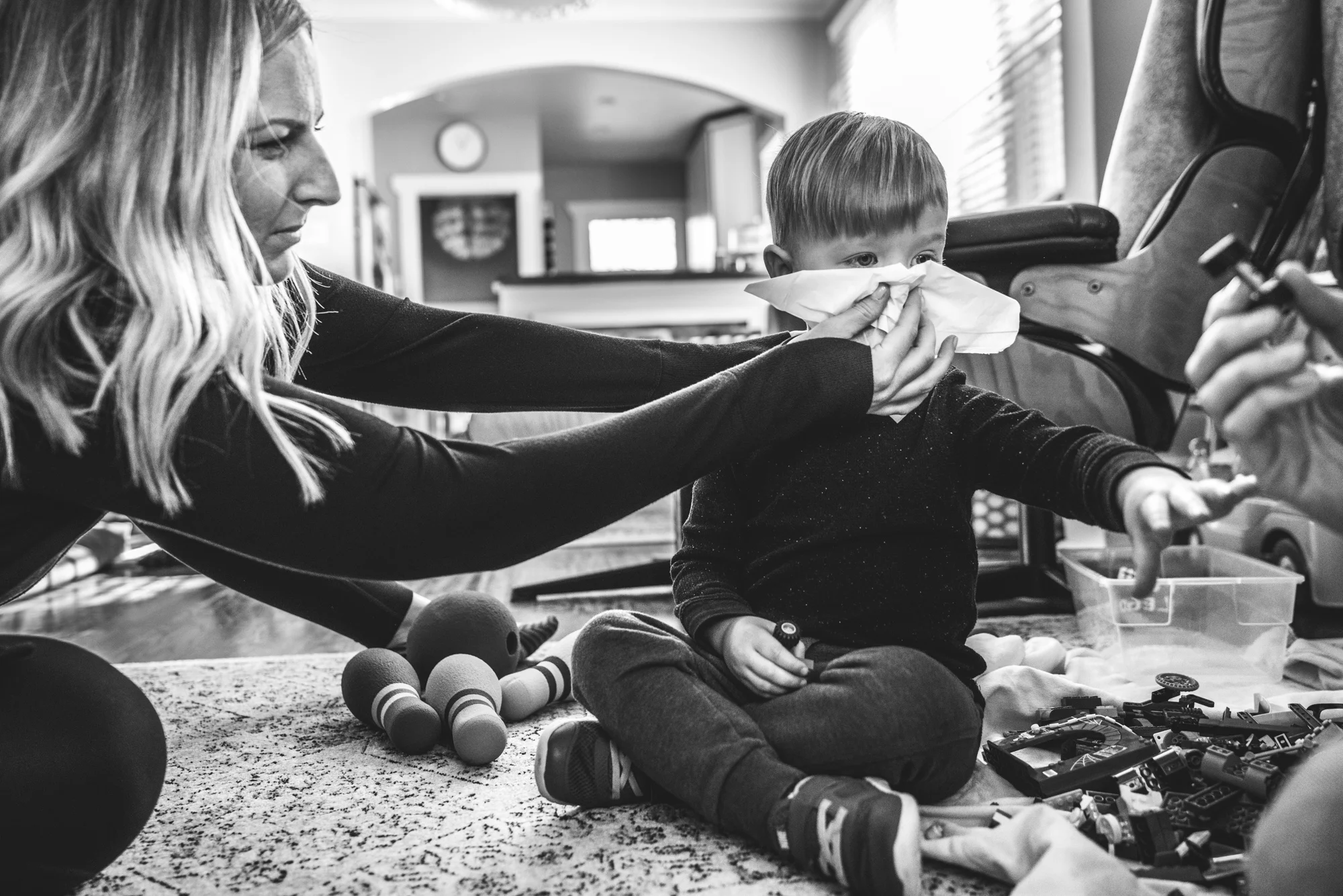 Black and white photo of a little boy reaching for a lego as his mother wipes his nose during their documentary family photography session at their home in Park Hill, Colorado.