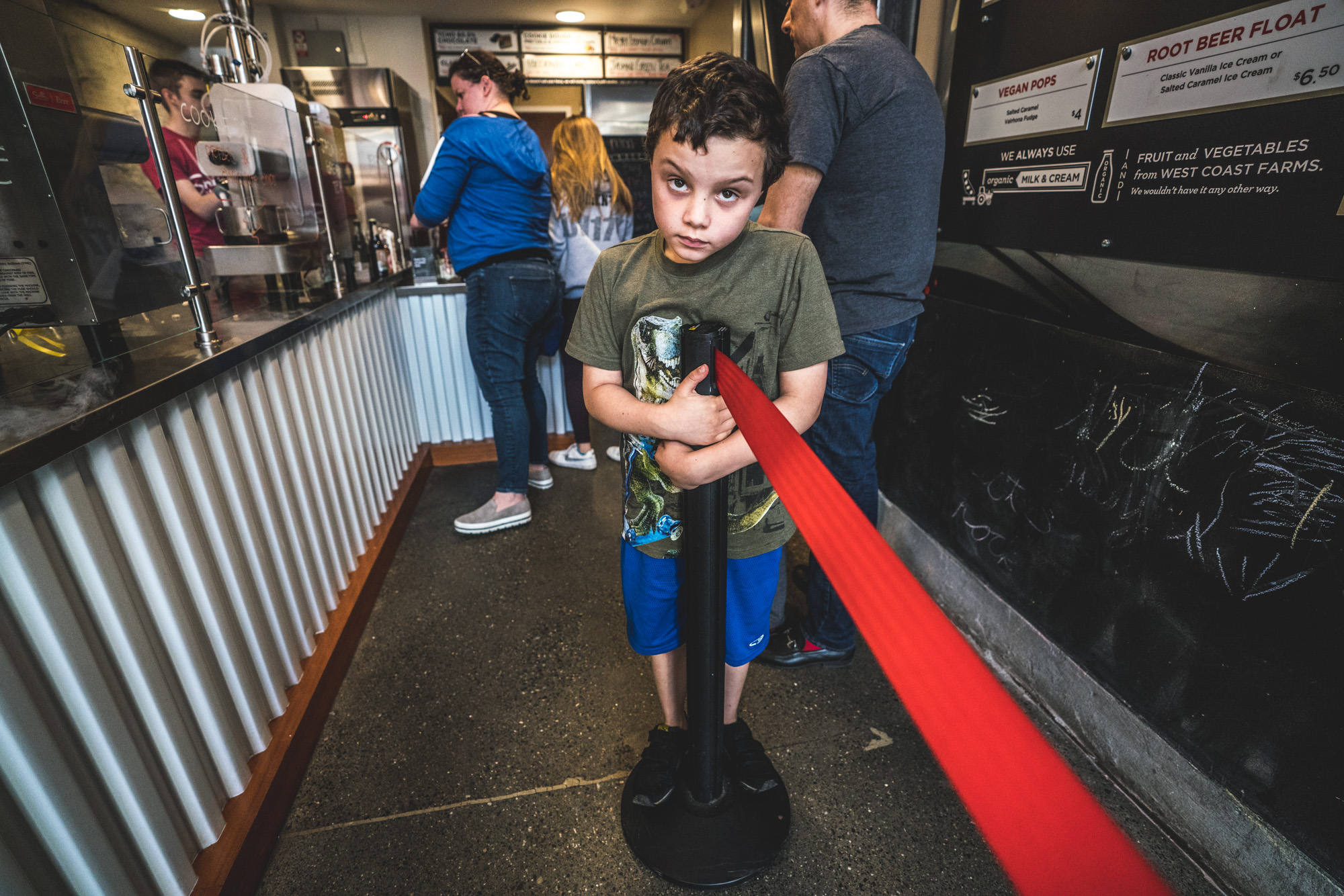 Color photo of a little boy in an ice cream shop, staring straight to camera and standing on a pylon (like he's not supposed to be)