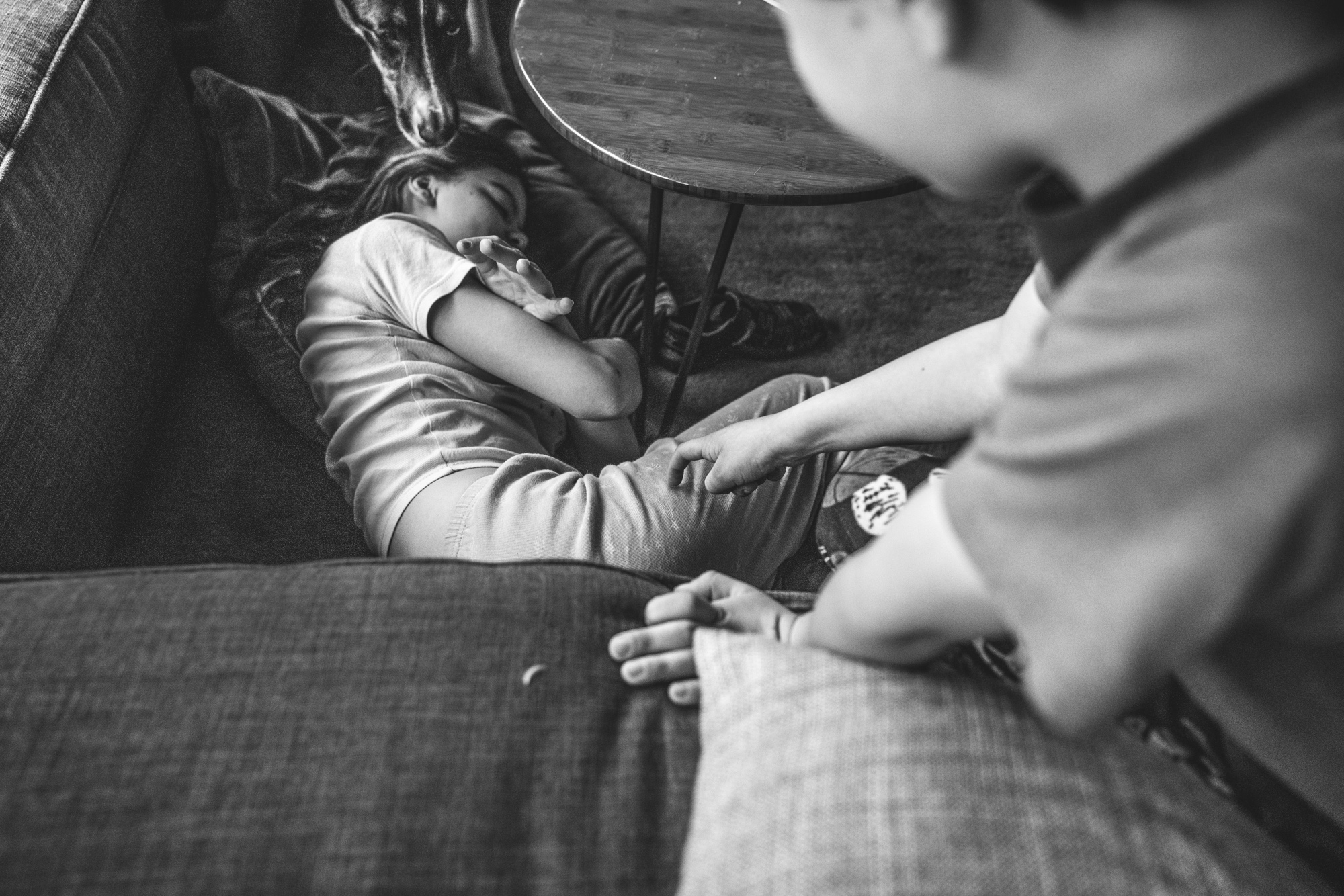 Black and white photo of a young girl lying next to the couch as her younger brother and dog try to wake her up; taken during her family's full day photography session