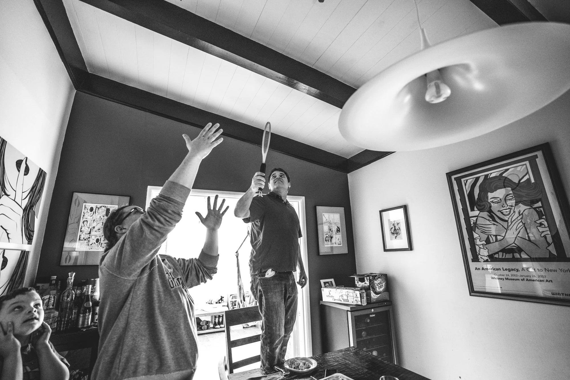 Black and white photo of a mother pointing to the ceiling as her husband stands on a chair holding an electric fly zapper, searching for the fly to kill. Photo taken during their family photography session in Oakland, California.