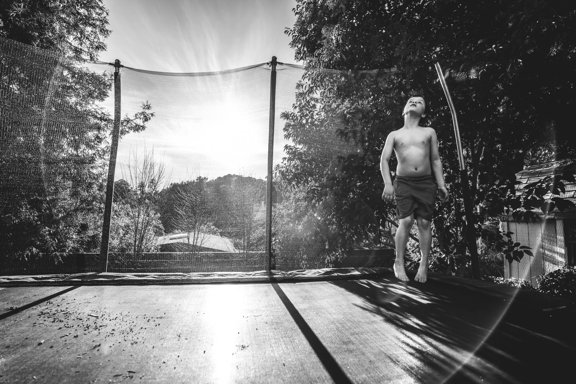 Wide black and white shot of a little boy jumping on his trampoline in his backyard in Oakland, CA