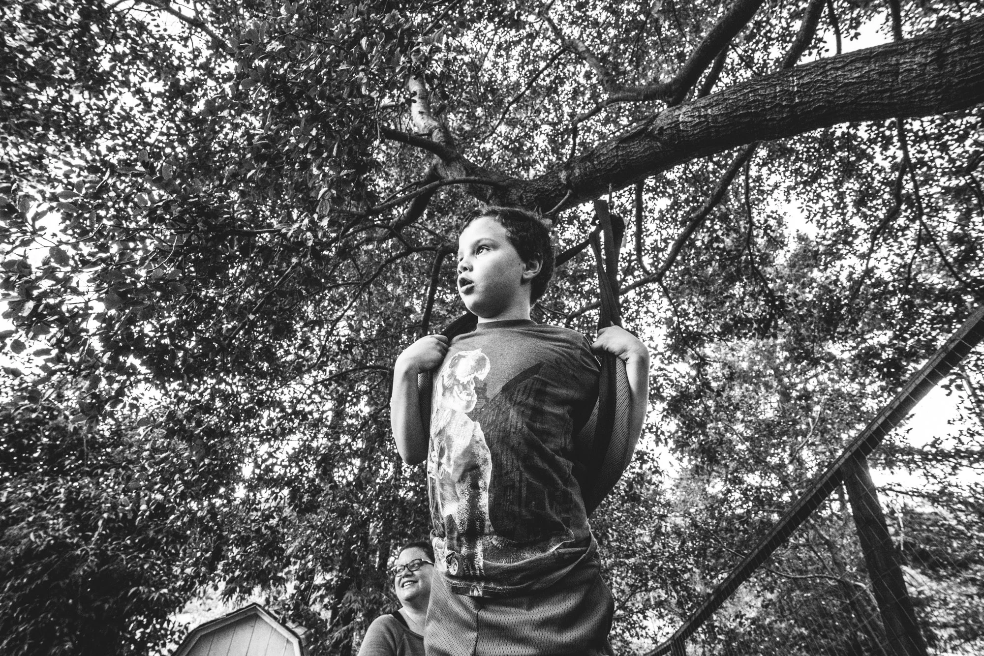 Black and white photo of a little boy standing up and holding onto his swing with a background composed entirely of the leaves of the many trees in his back yard; taken during his full day in the life photography session