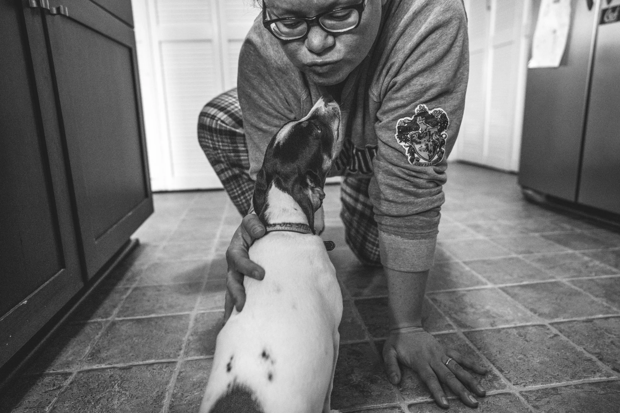 Black and white photo of a mother leaning down on the kitchen floor to kiss her small black and white dog at their home in Oakland during their documentary photography photo session