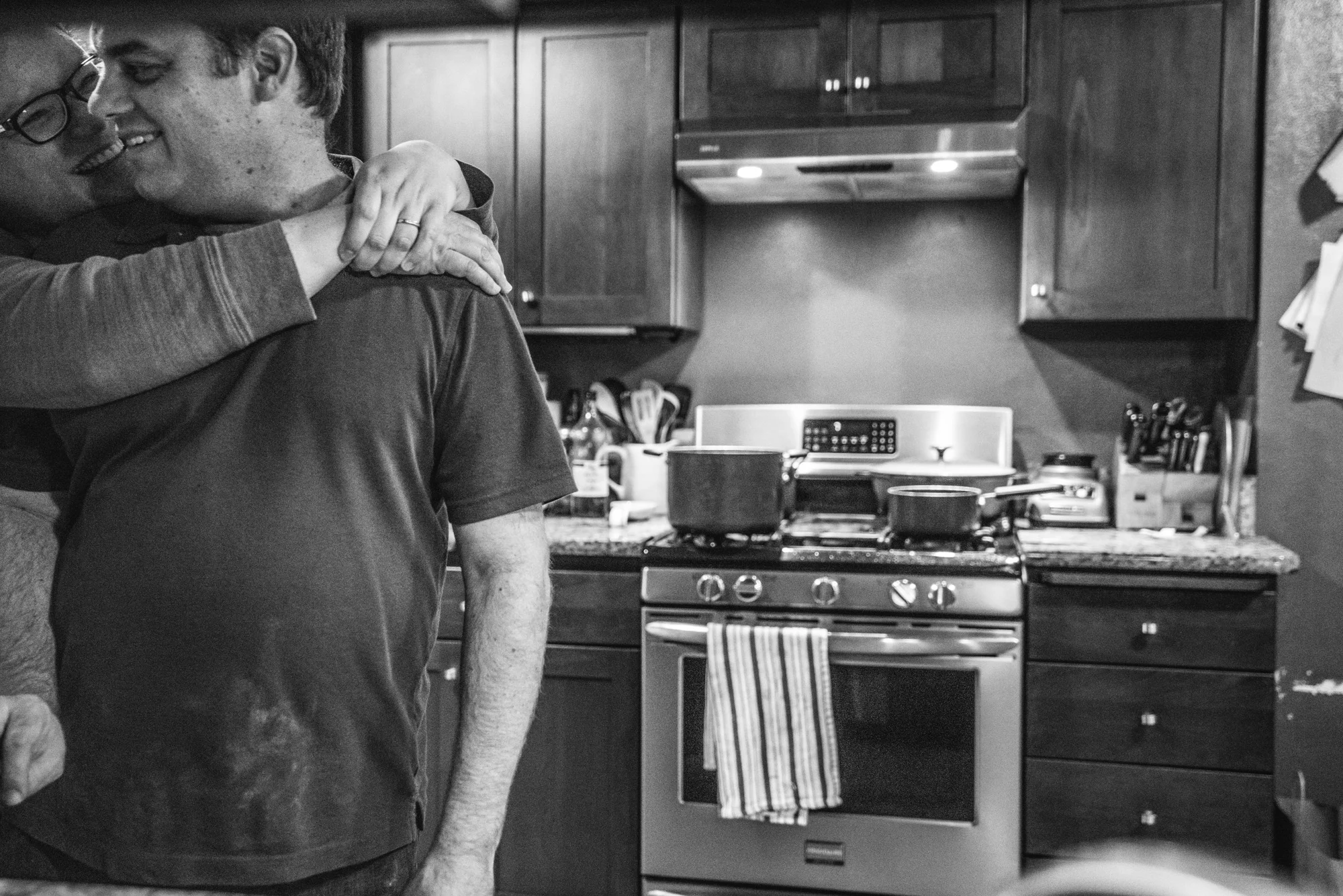 Black and white photo of a husband and wife in their kitchen, facing the camera with the husband in front and the wife behind him with her hands thrown around his neck, both smiling at each other. Taken at their home in Oakland, California during their documentary photography session