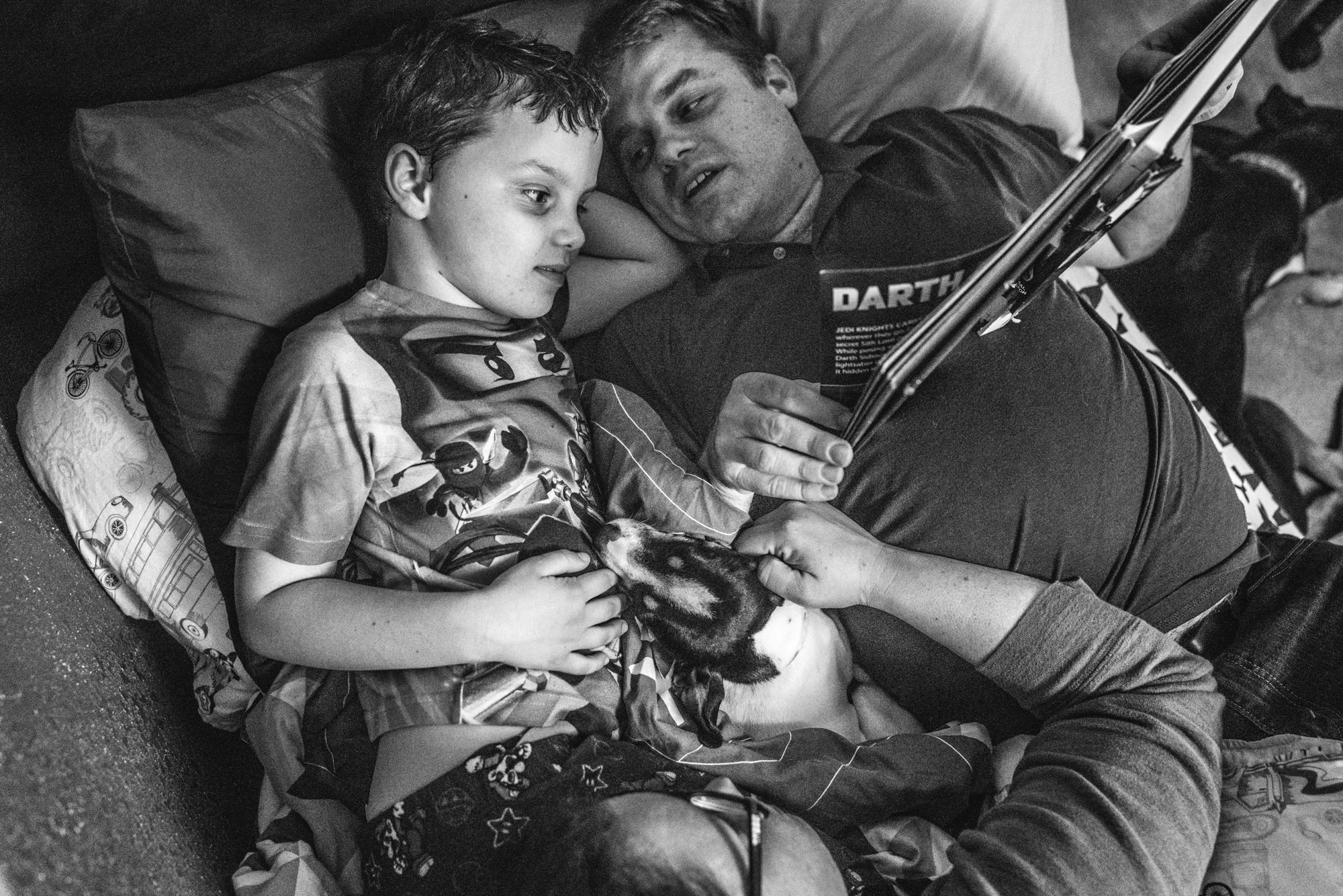 Black and white photo of a father and son reading a star war's book during story time while the mother and their little dog lie at their feet, taken during their family photography session in their home in Oakland, CA