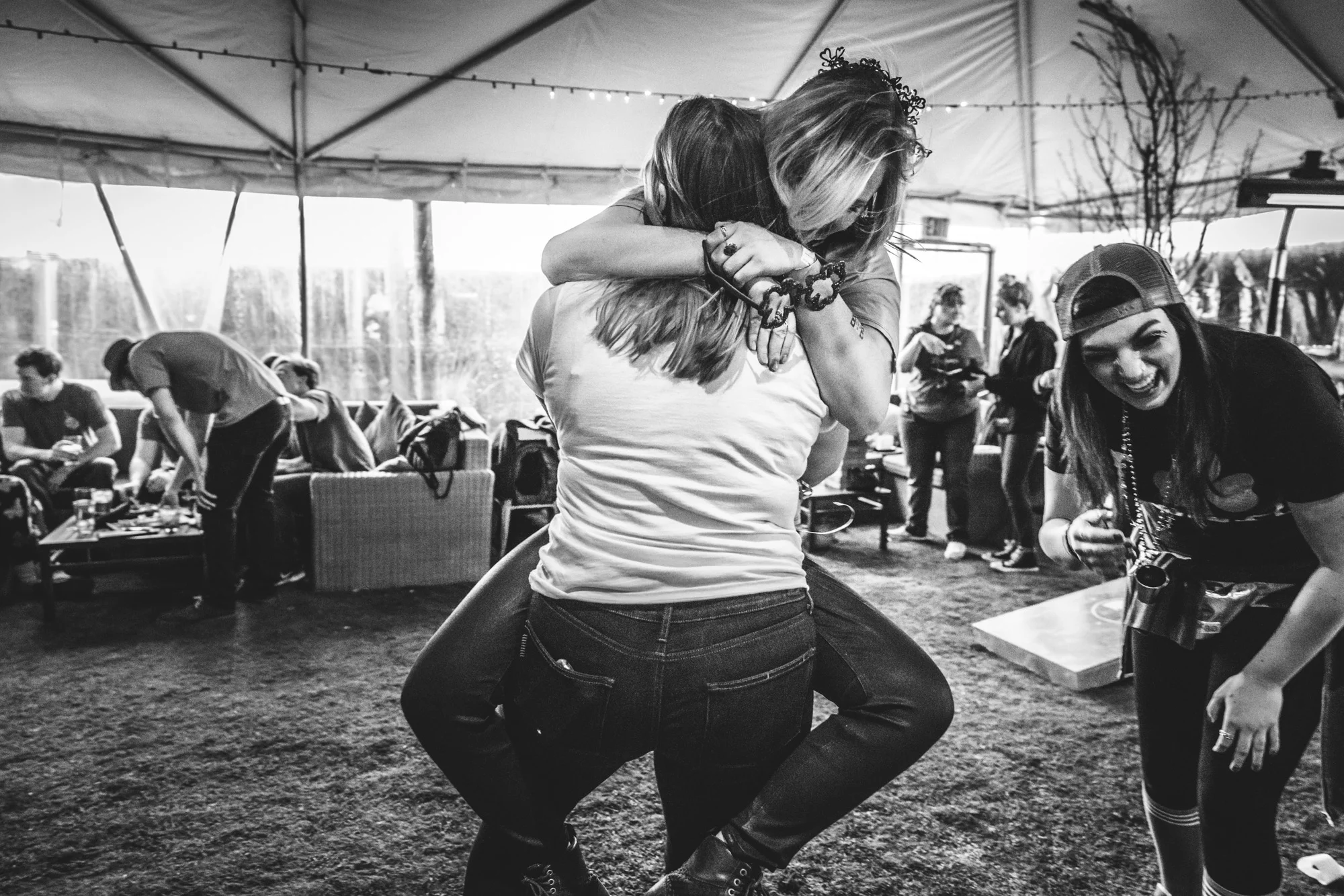 Black and white photo of a group of people in a tent outside of the ViewHouse in downtown Littleton, Colorado. One woman jumps up on another as their friend standing to the side of them laughs. Taken during the Autism Society of Colorado Shamrock Stumble in March 2018.