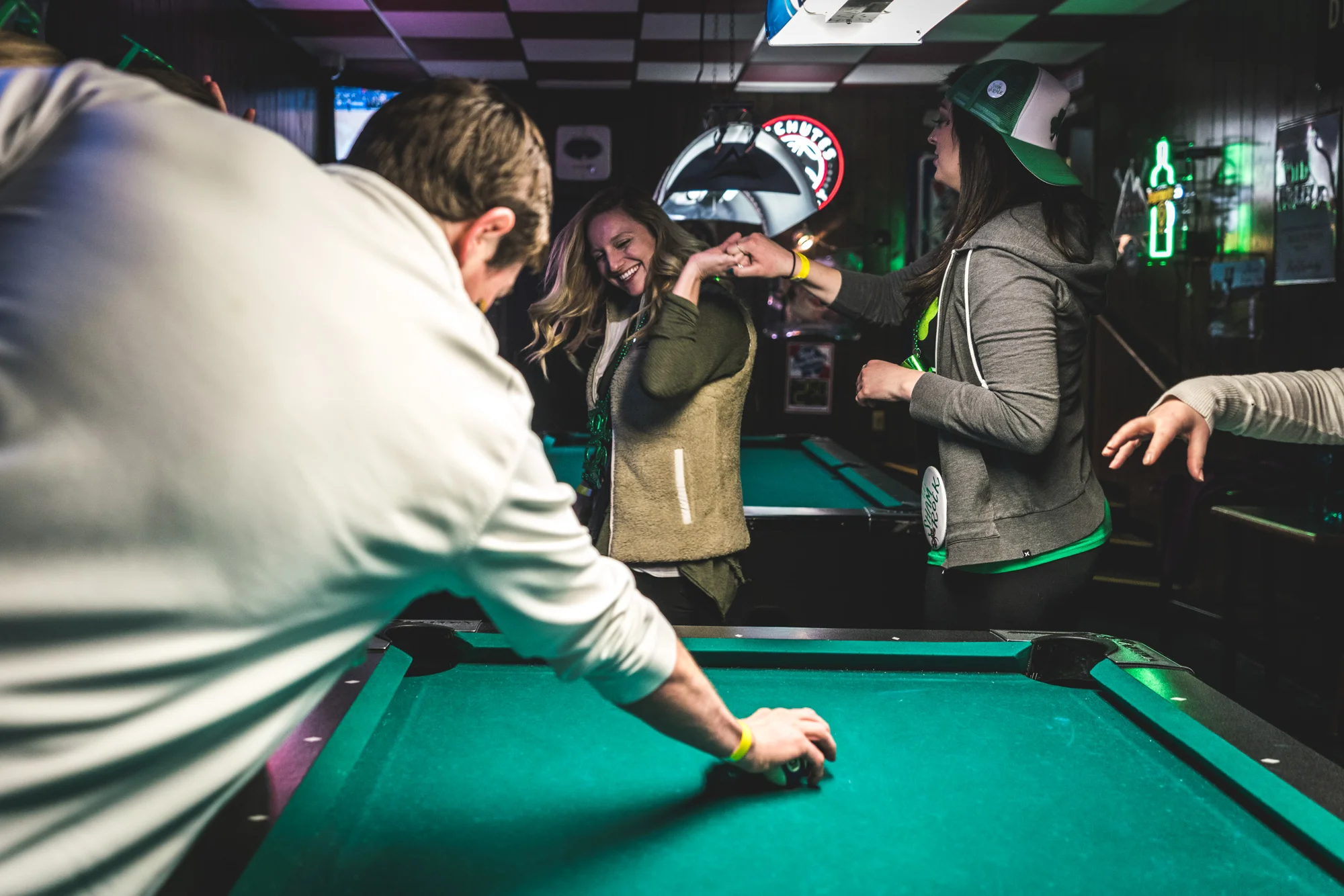 Color photo of two woman dancing near a pool table as a man stretches out to grab a ball on the pool table. Taken at the Olde Towne Tavern in Littleton, CO for the ASC Shamrock Stumble fundraiser event.