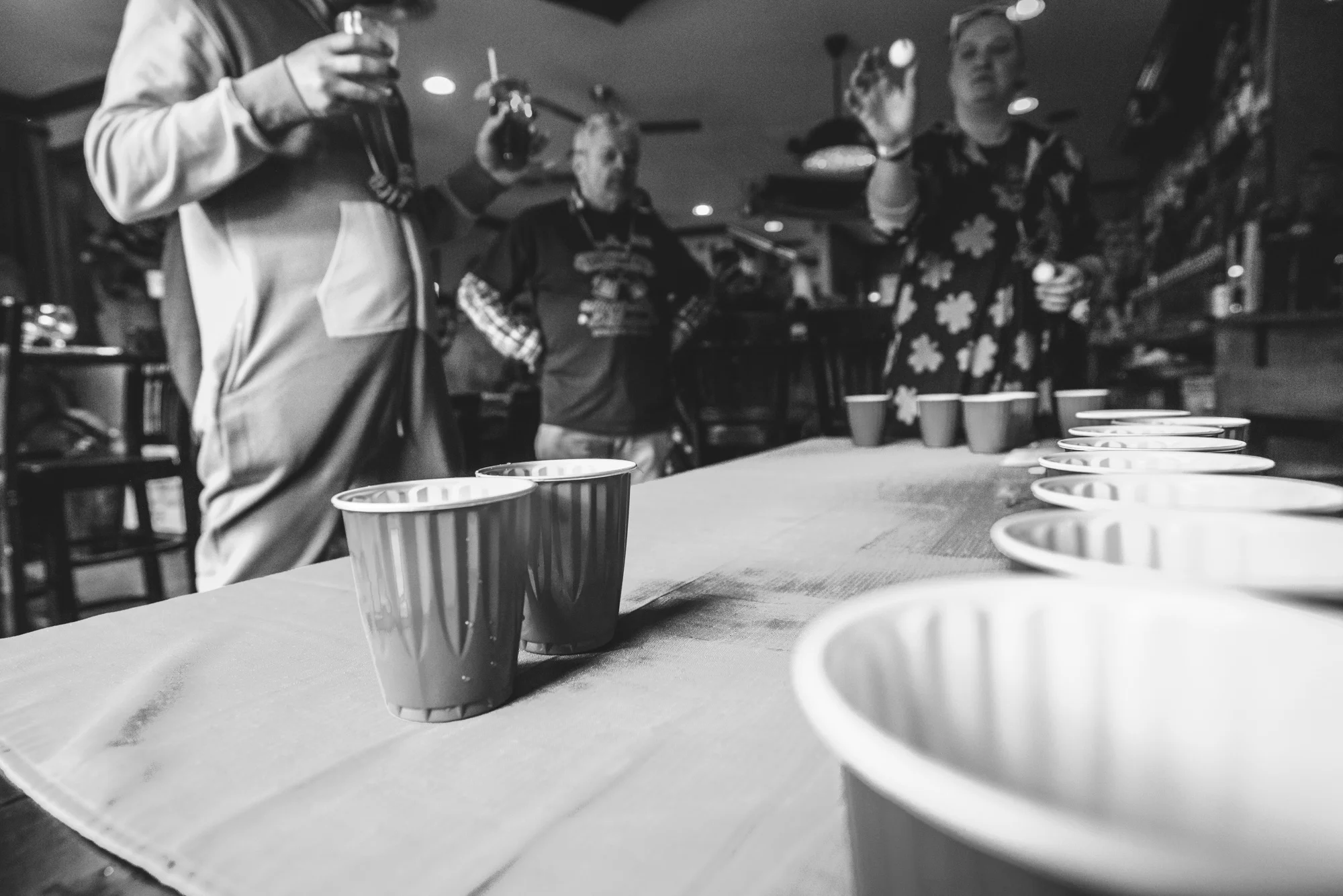 Black and white photo of a woman taking careful aim with a ball at two solo cups on a table as she plays Beer Pong at Jake's Brew Bar during the documentary photography session of the ASC Shamrock Stumble. 