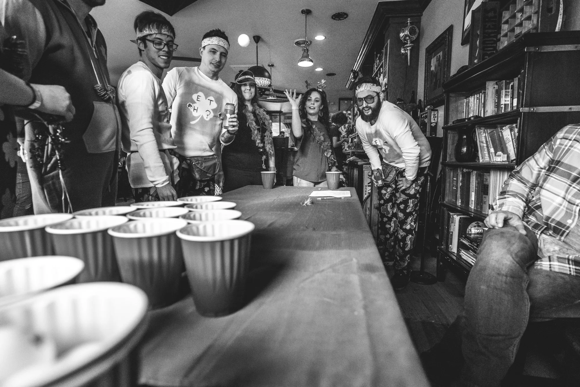 Black and white photo of a woman throwing a ball at solo cups during a game of Beer Pong as a bunch of people stand around and watch. Taken during the documentary photography event session of the Autism Society of Colorado's Shamrock Stumble fundraiser at Jake's Brew Bar.