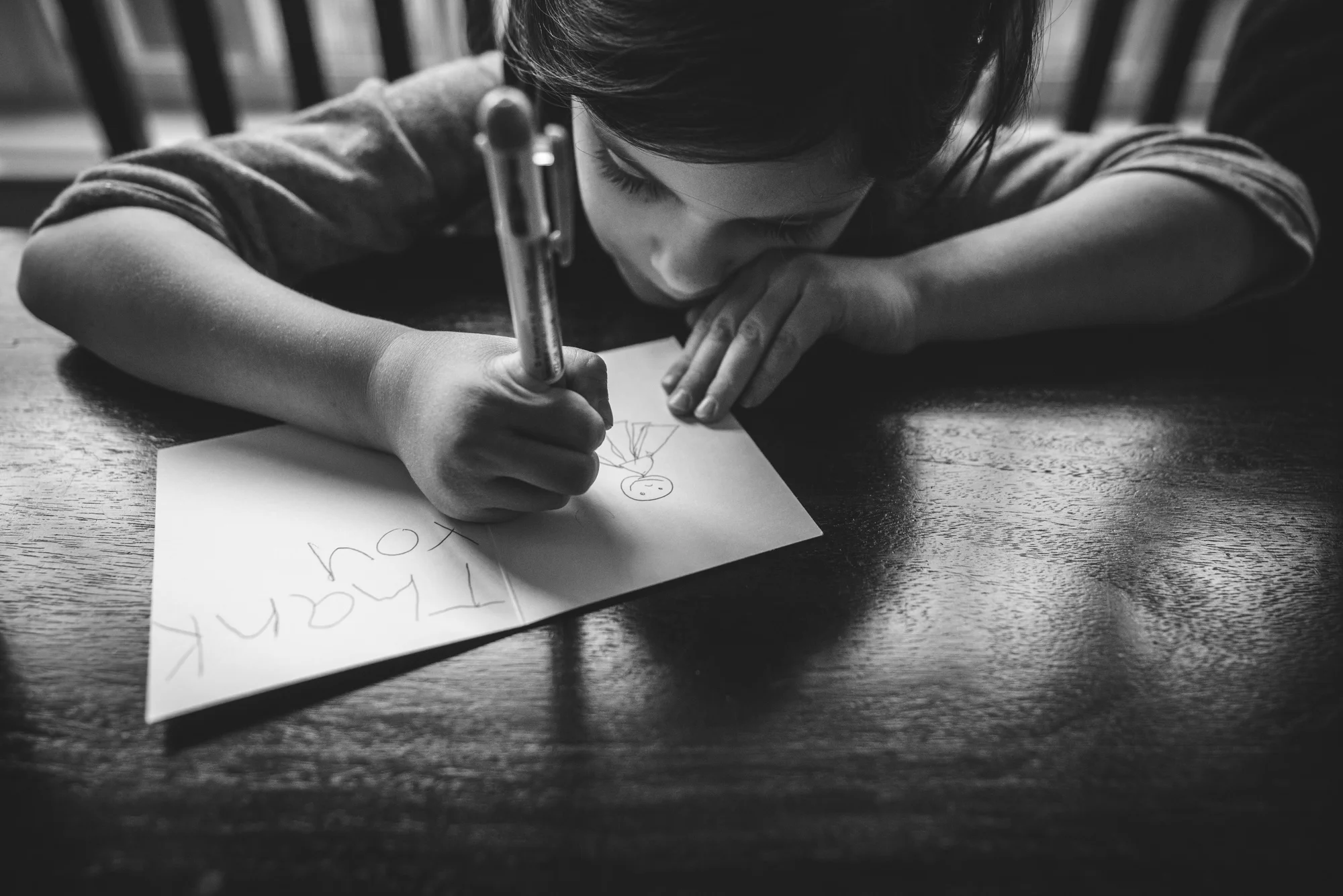 Black and white photo of a sweet little girl writing out a thank-you card and drawing a picture of a ballerina. Taken at her kitchen table in Denver, Colorado during her family's at-home photography session.