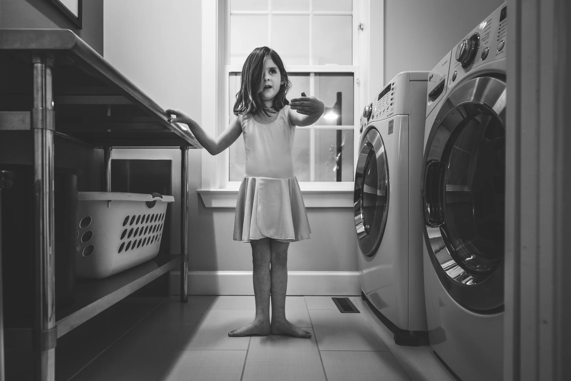 Black and white photo of a 6-year-old girl in her ballet costume doing one of the ballet positions while standing in her laundry room in her home in Stapleton, Colorado. Taken during her family's documentary photography session.