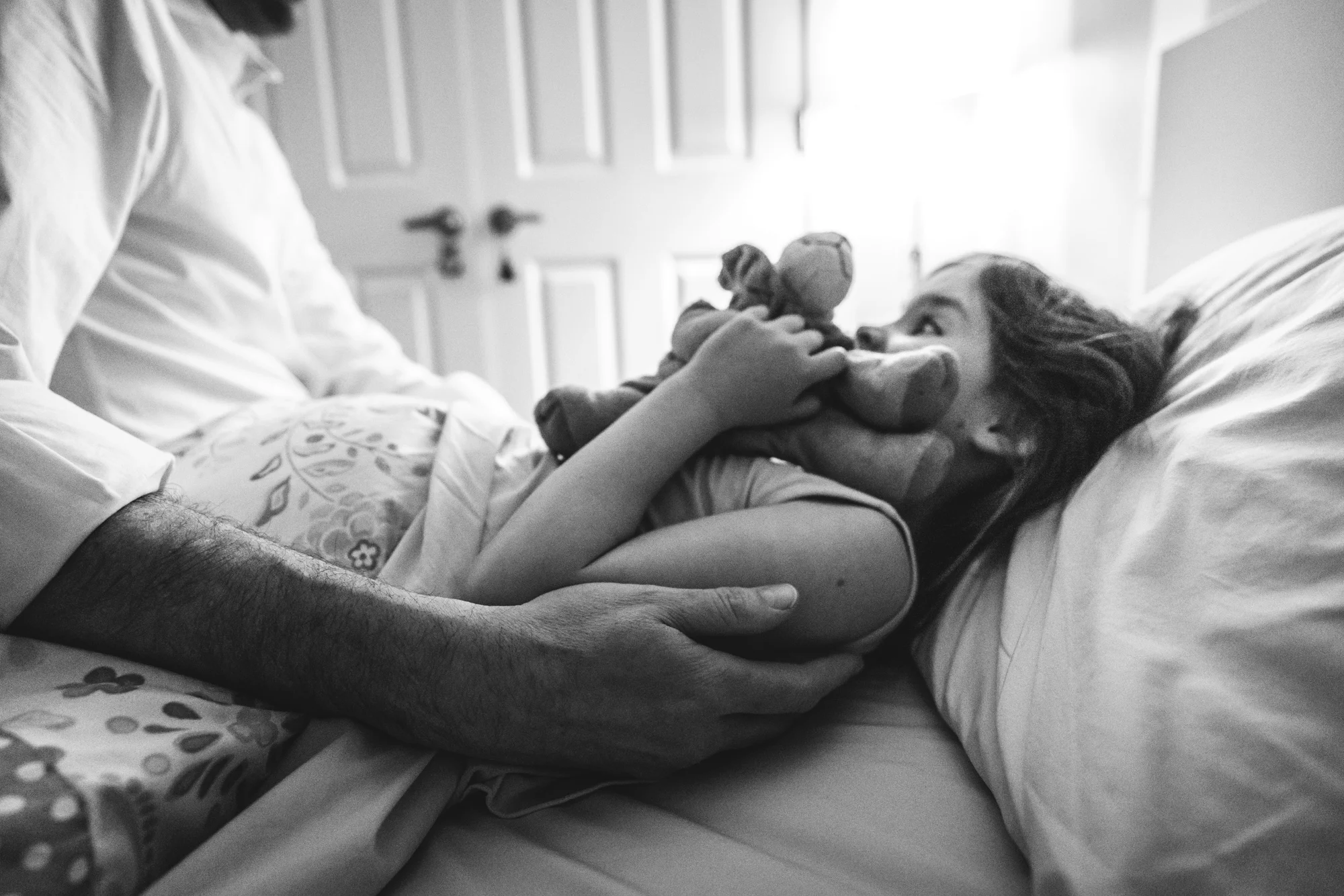 Black and white photo of a father tucking his young daughter into bed as she holds her stuffed lion close to her. 