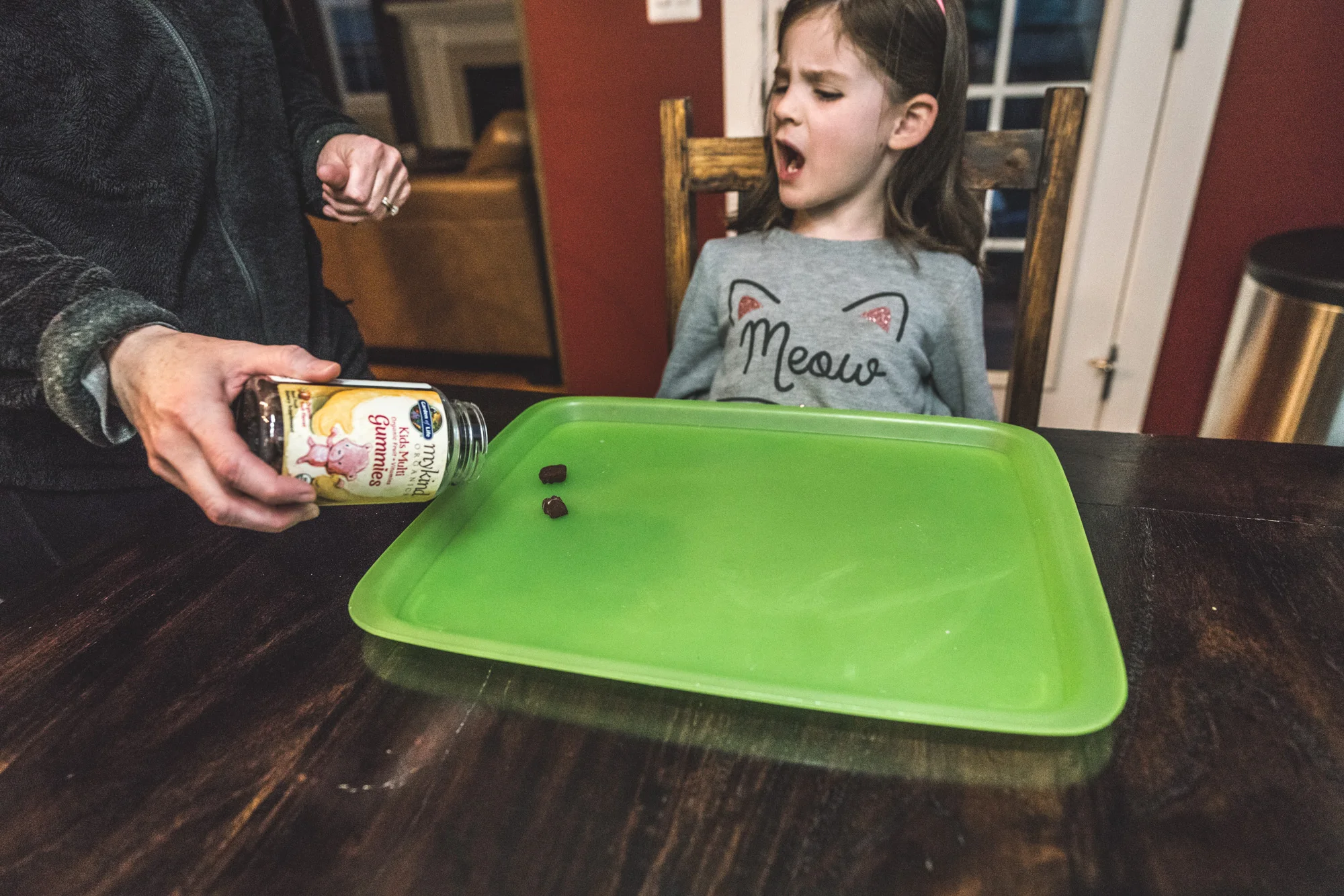 Color photo of a mother pouring vitamins out onto a green tray for her little girl who is sitting at the table wearing a cat-ear headband and a "meow" shirt. Taken at their home in Denver, Colorado during their at-home photography session.
