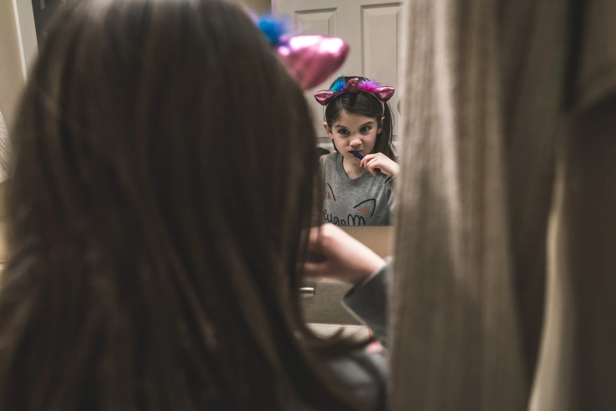 Color photo of a little girl wearing a pink unicorn headband while brushing her teeth intently. Photo taken in her home in Denver, Colorado.