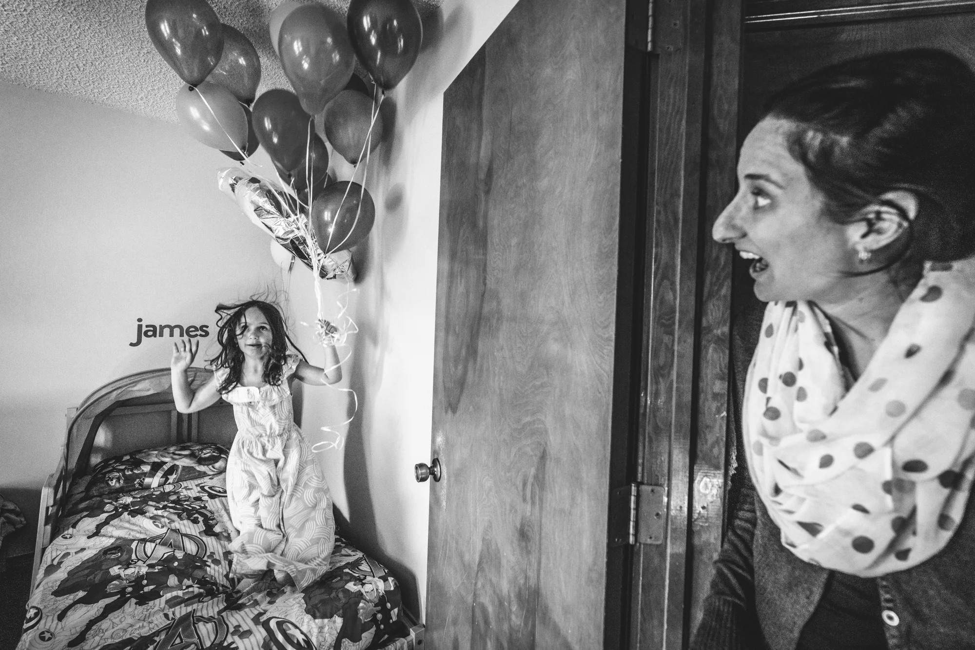 Black and white photo of a little girl at a birthday party, jumping on a bed while holding a bunch of balloons while her mother looks on with a shocked expression on her face, taken in Denver, Colorado