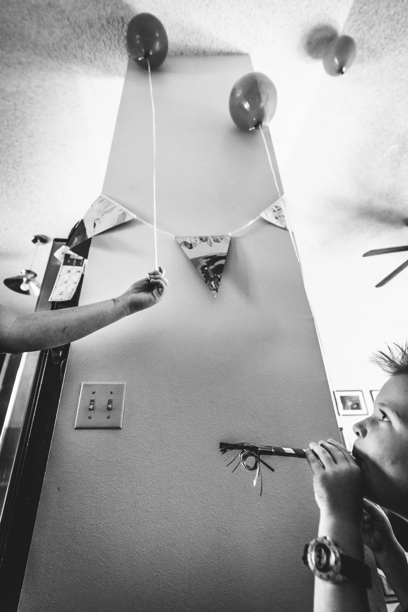 Black and white photo of a woman's arm retrieving a balloon for her son, who's blowing a party blower