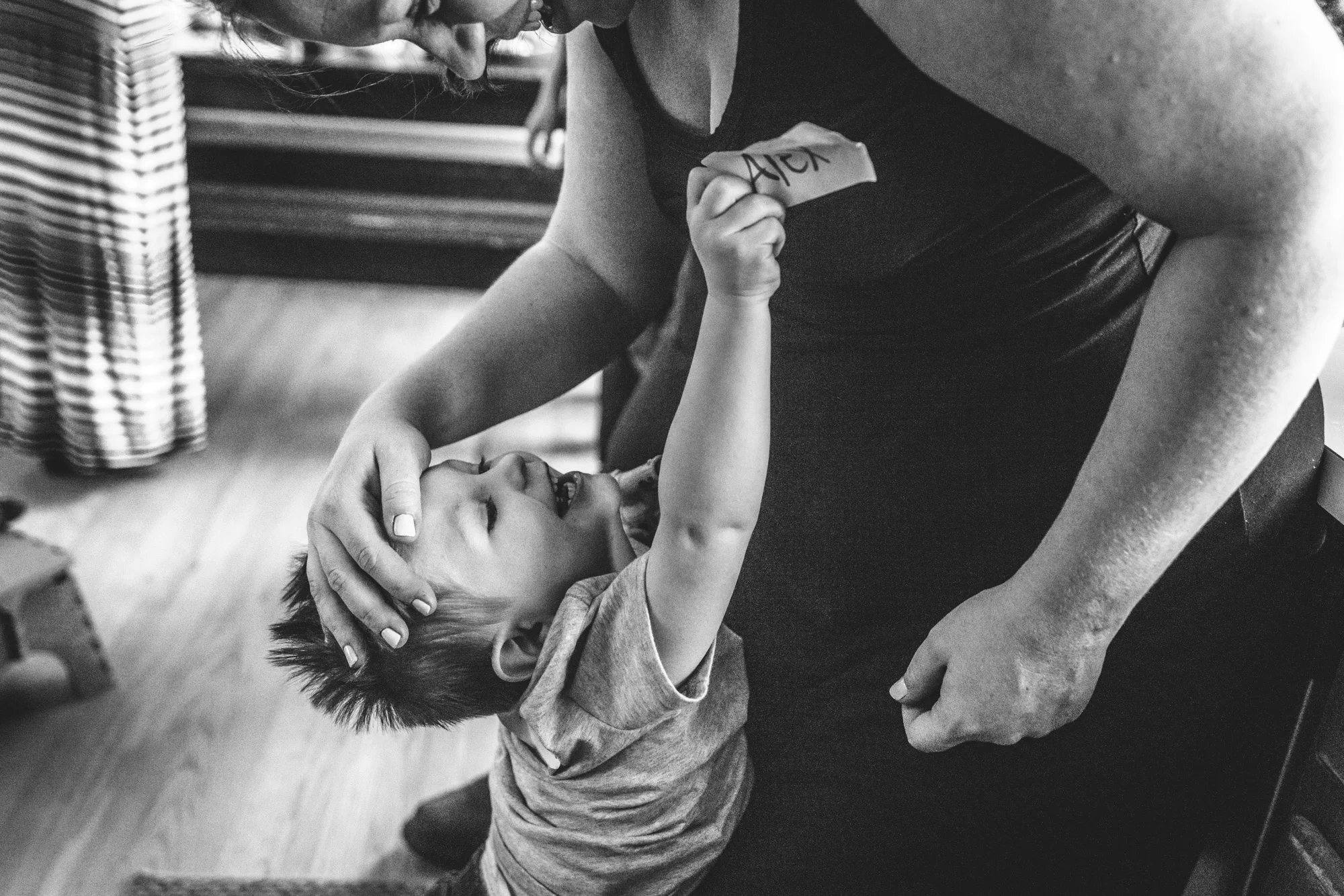 Black and white photo of a little boy trying to steal his mom's name tag; taken in Aurora, Colorado during an at home birthday party