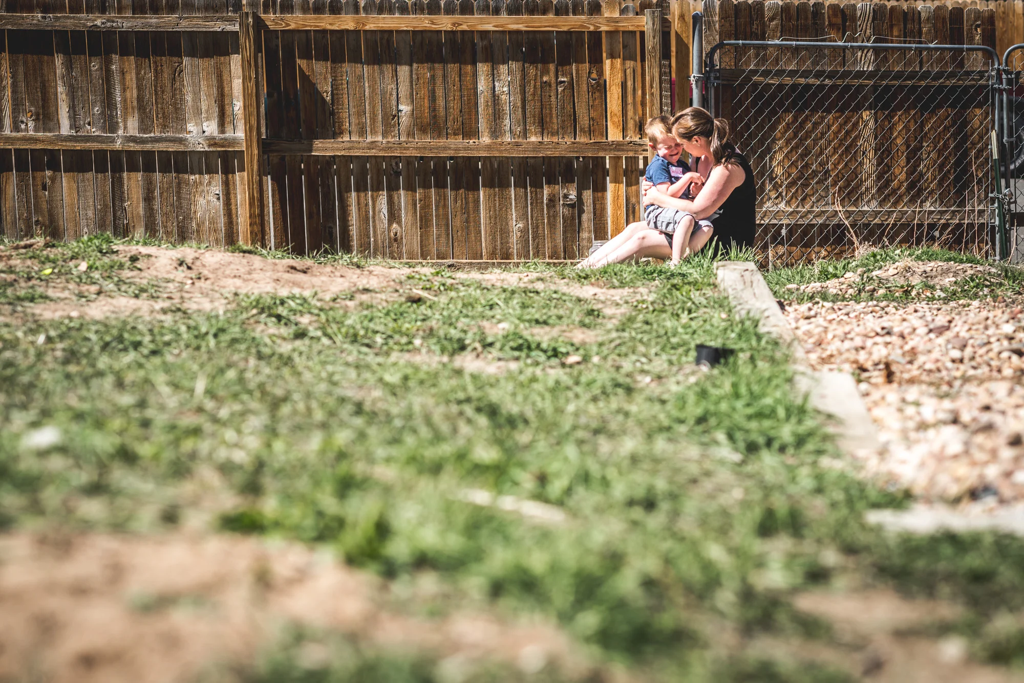 Color photo of a little boy sitting and crying on his mother's lap in their back yard as she comforts him