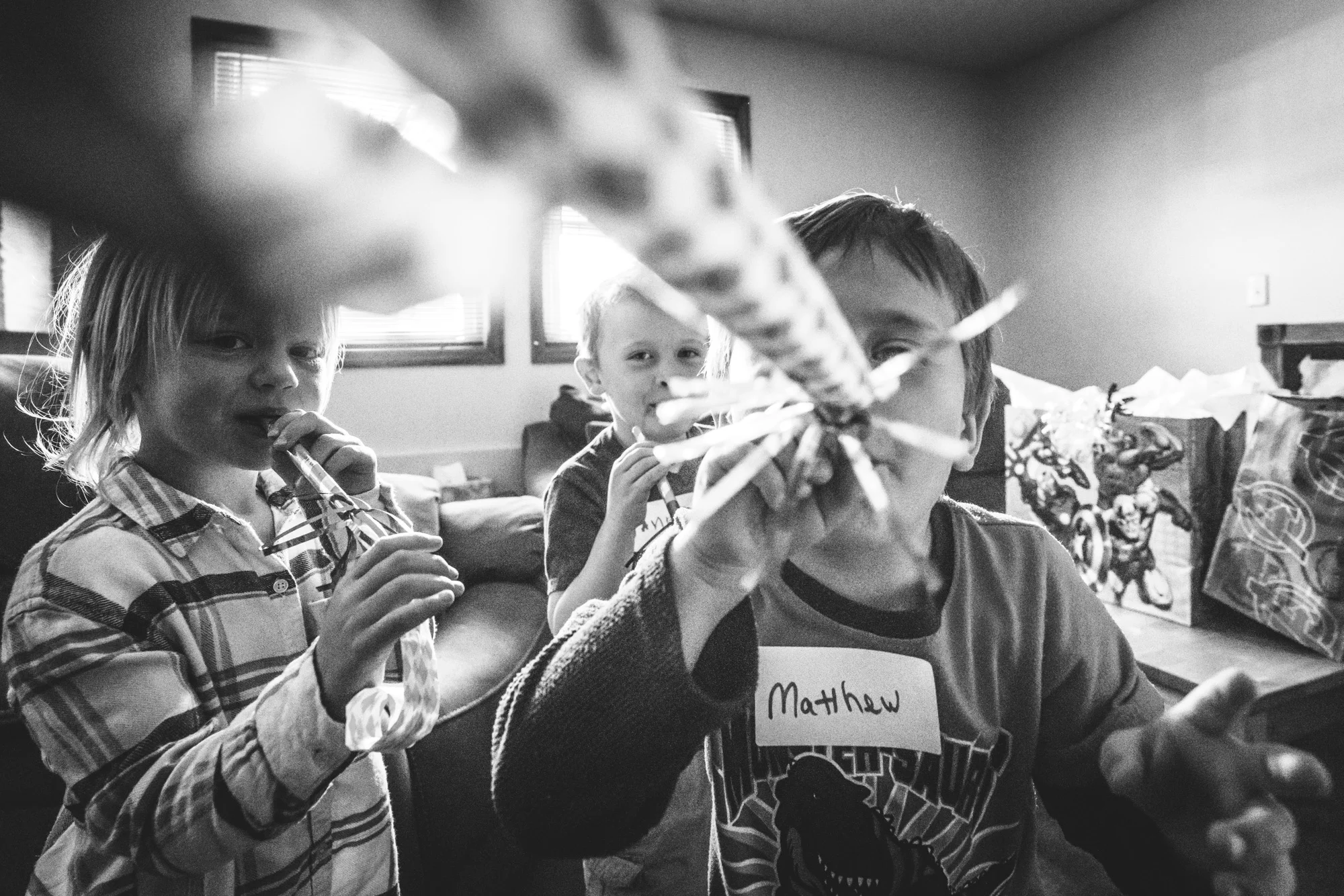 Black and white photo of a little boy blowing a party blower directly at camera while his two friends watch