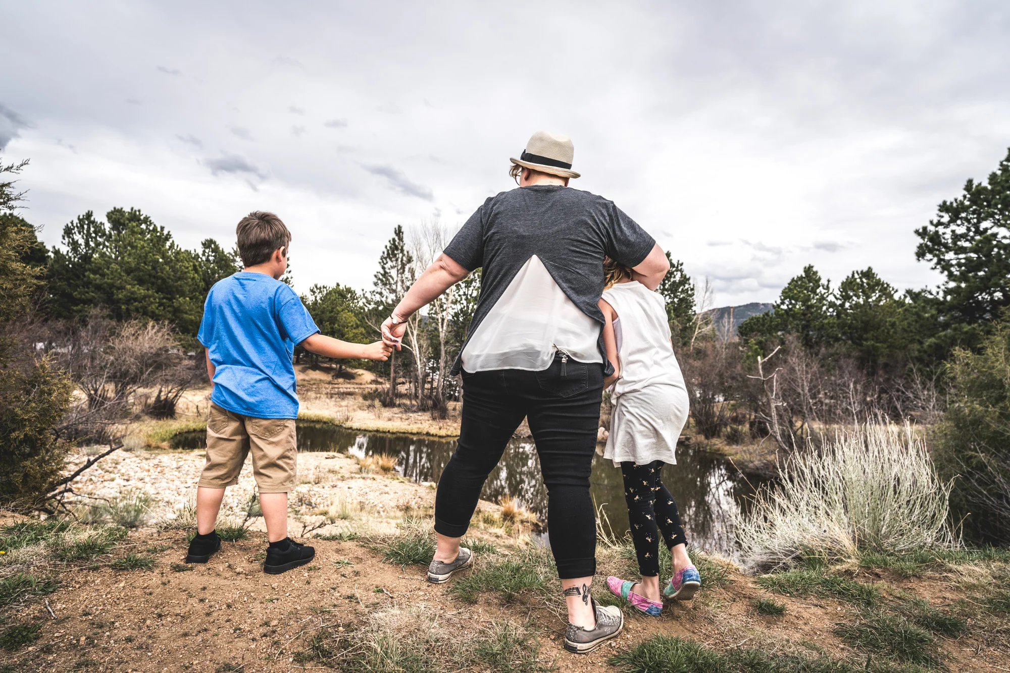 Color image of a woman holding her young daughter under one arm and holding her young son's hand in the other as they look upon a pond. 