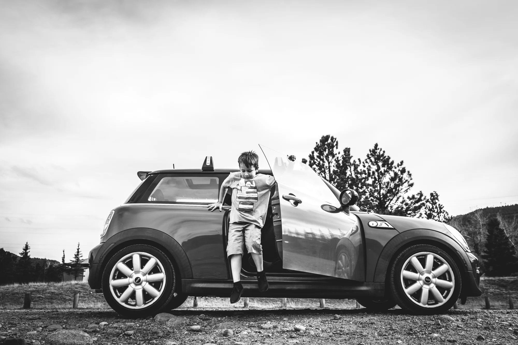 Black and white photo of a boy mid-air as he jumps out of a mini cooper. Taken during his mini adventure photo session in Estes Park, Colorado.
