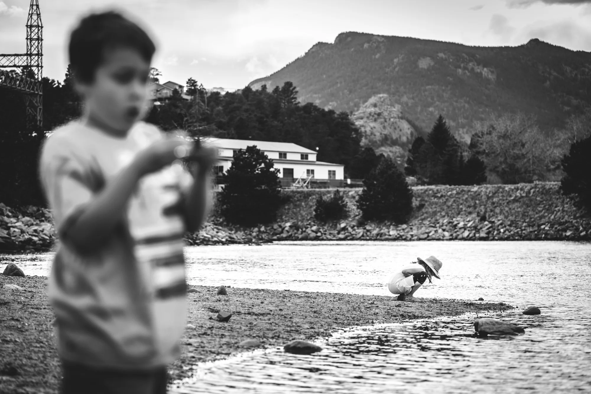 Black and white photo of a little girl bent down, inspecting something in the rocky, sandy beach of a lake in Estes Park, Colorado as her brother in the foreground looks at something in his hand.