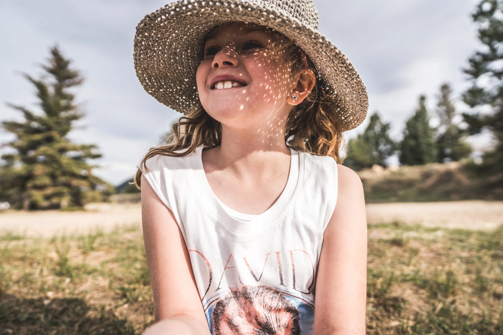 Color environmental portrait of a blond, curly-haired little girl smiling as she wears a straw hat that creates dapples of light on her face as the sun shines through it. Taken during her family documentary photo session in Estes Park, Colorado.