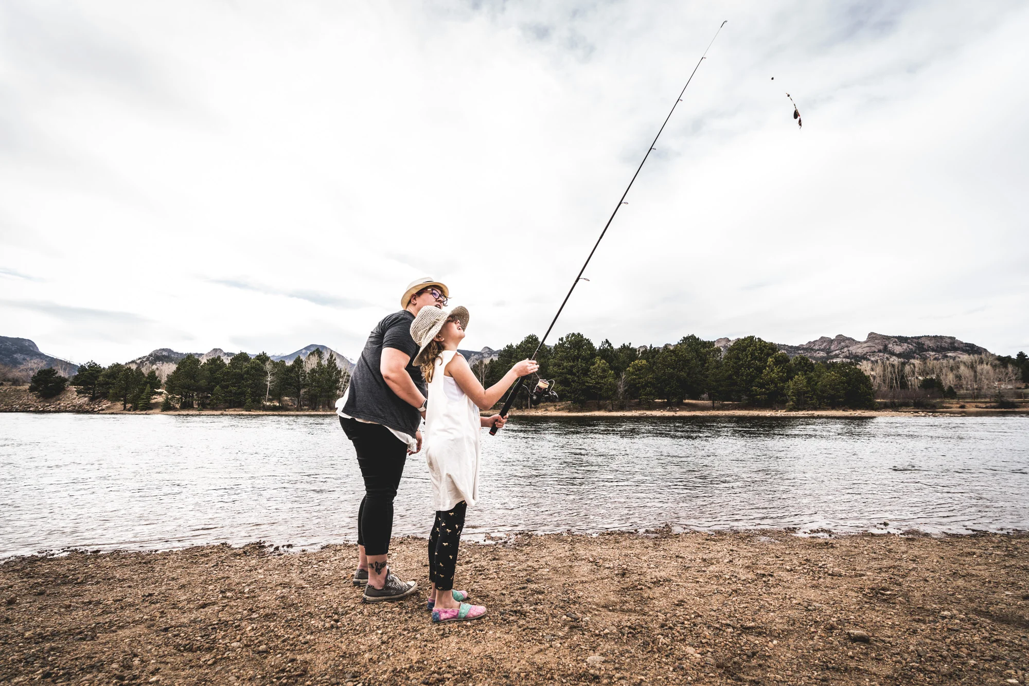 Color photo of a mother and daughter looking up to the top of the fishing pole held by the young girl. Taken in front of a lake in Estes Park, Colorado.