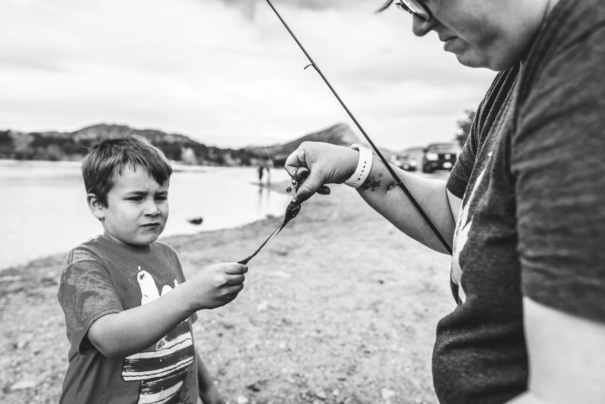 Black and white photo of a mother holding onto a fishing rod covered in algae as her young son attempts to pull the algae off. Taken by a lake in Estes Park, CO.