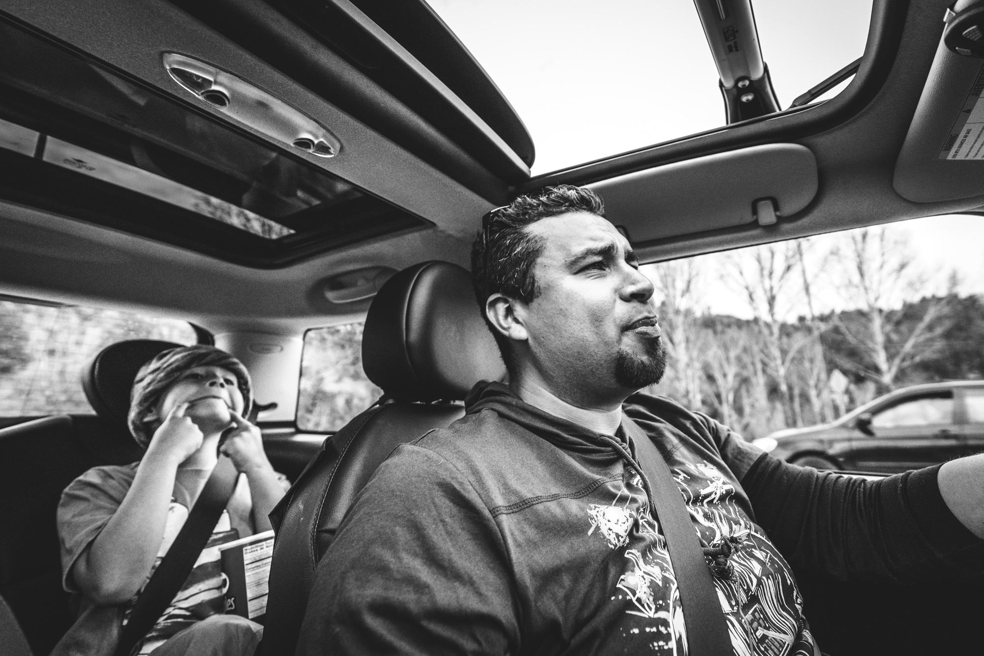 Black and white photo of a father driving a car as his son makes a funny face at him from the back seat. 