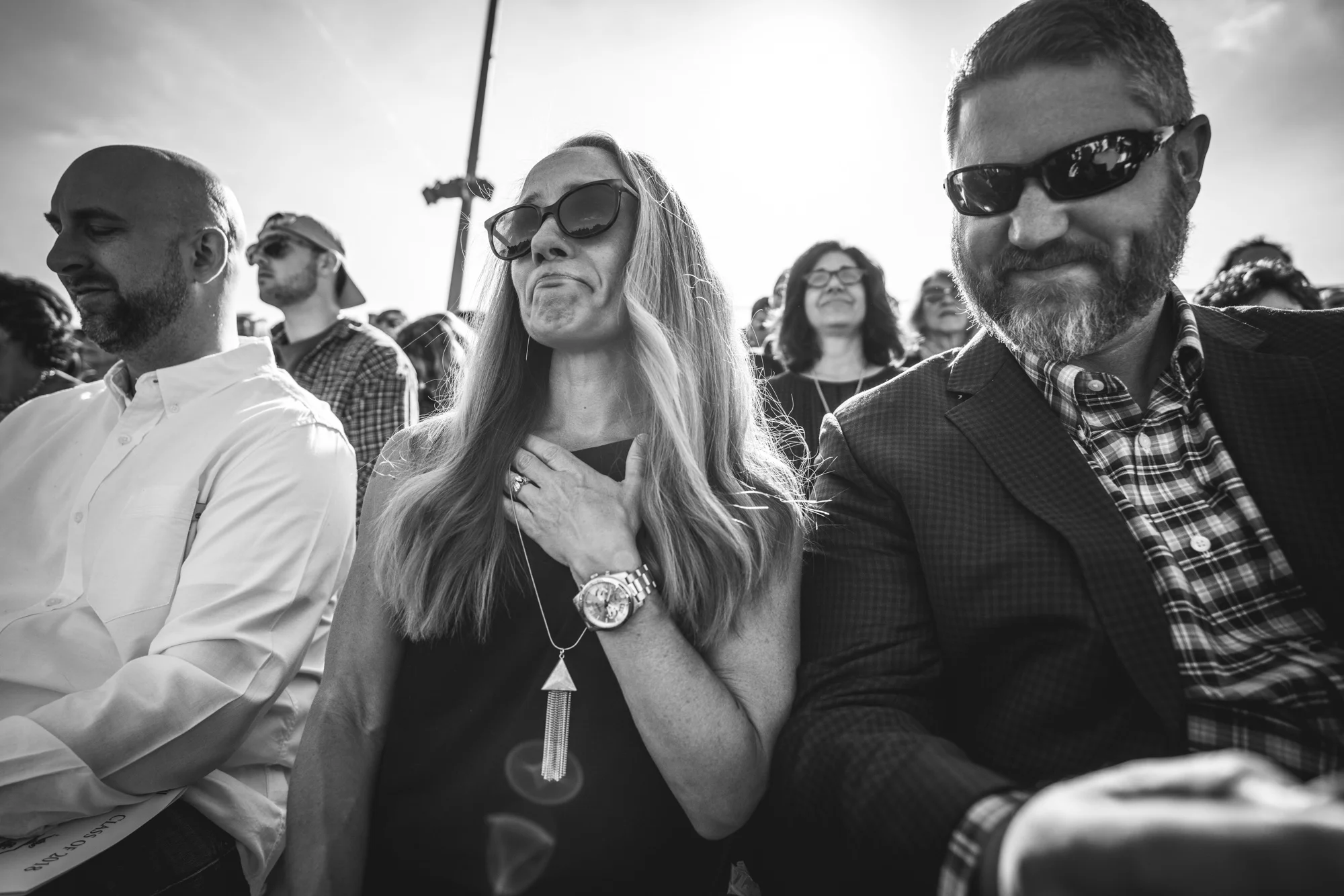 Black and white photo of a woman sitting in the bleachers at her son's graduation ceremony with her hand to her chest, crying tears of joy.