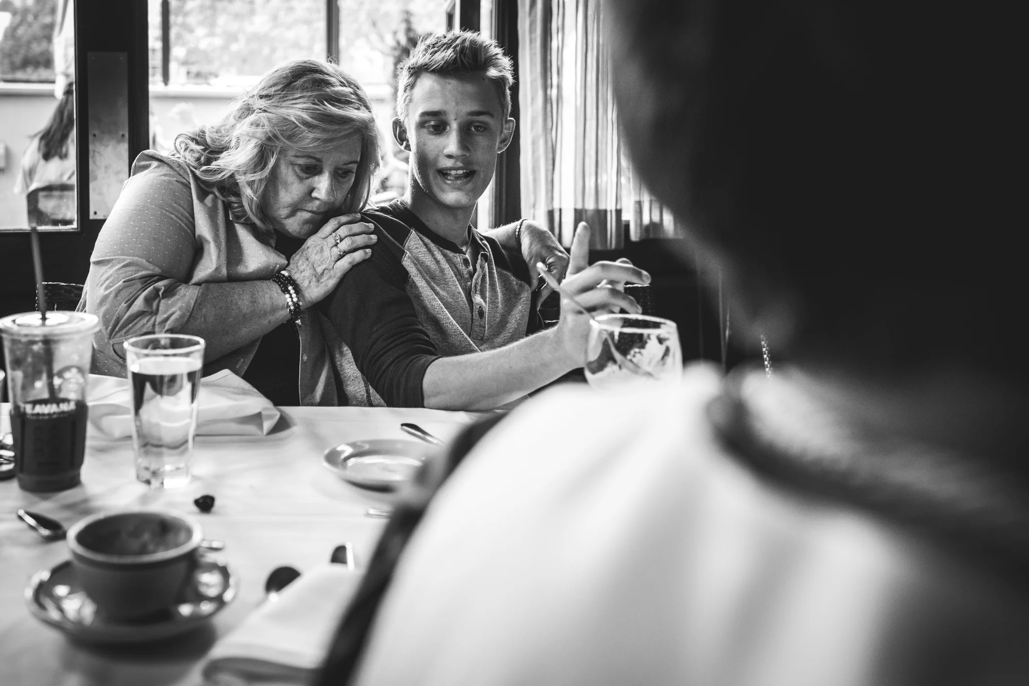 Black and white photo of a grandmother and grandson at a restaurant. The teenage grandson talks while his grandmother leans on and hugs him. Taken during a documentary event photography session in Denver, Colorado. 