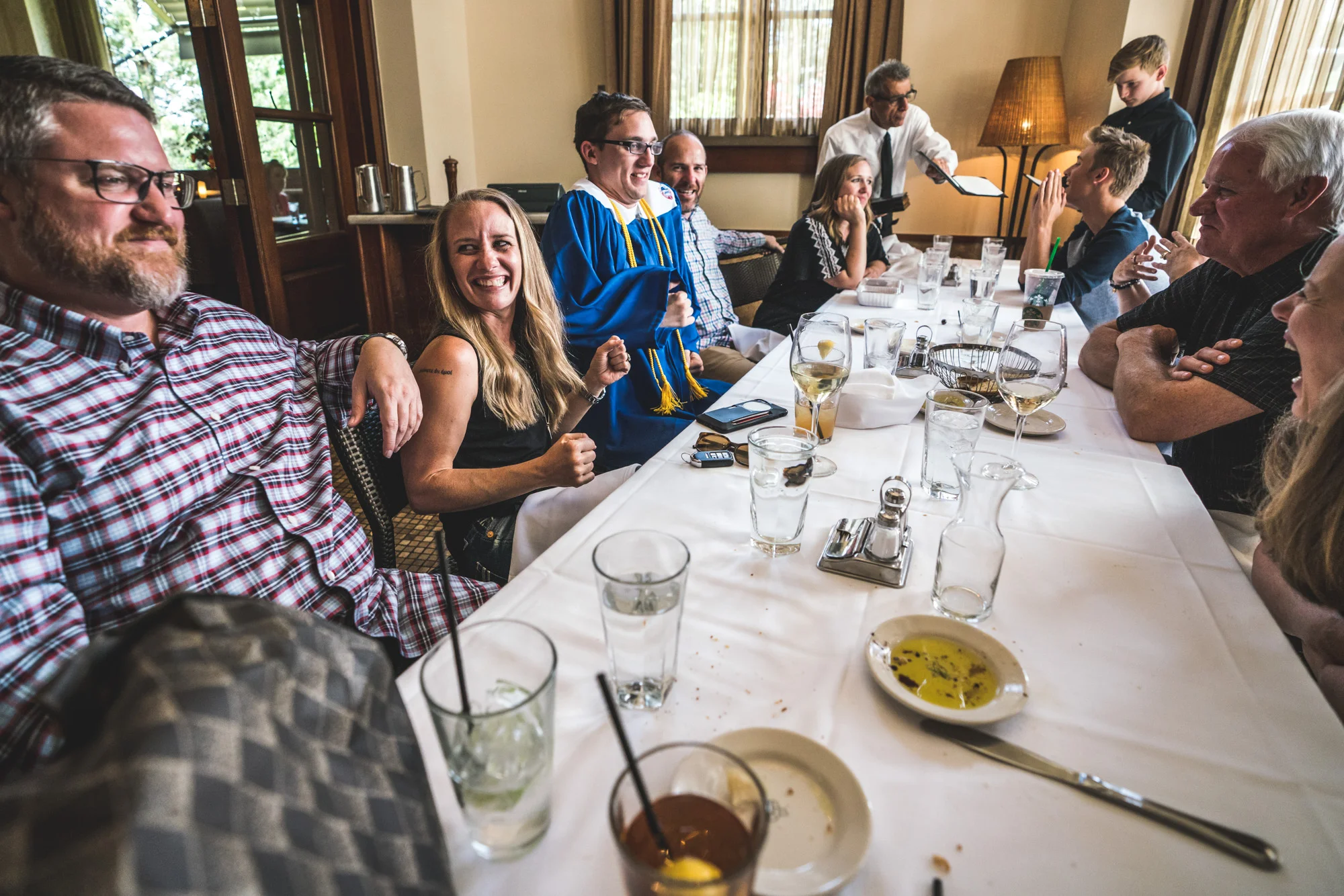 Color photo of a large family sitting around a table at a restaurant. Among the members is a teenager wearing his blue high school graduation cap and gown. Taken in Denver, CO.