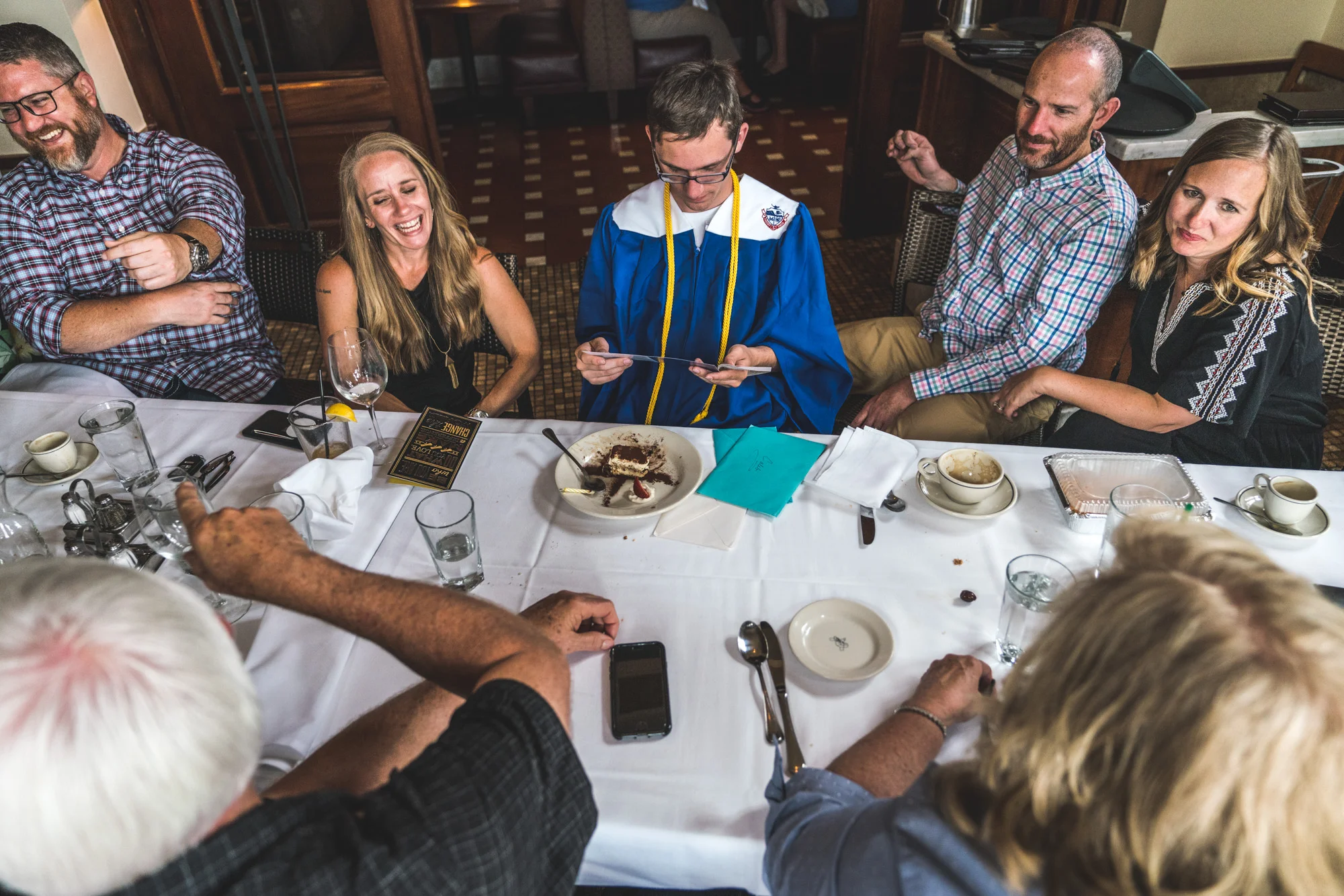 Color photo of a family sitting around the table in a restaurant with a teenager wearing his blue high school cap and gown after having graduated from high school.