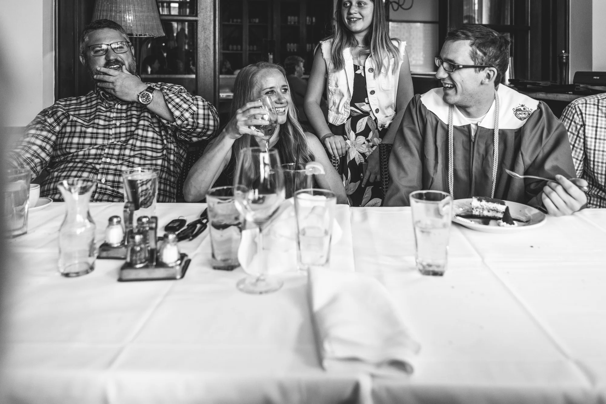 Black and white photo of a family at a restaurant together, the mother looking over and smiling at her teenage son, who has just graduation high school and is wearing his cap and gown.