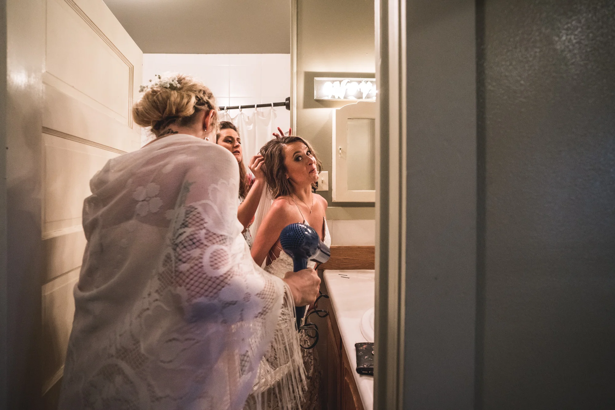 Color photo of a bride in a bathroom, amused as one woman attempts to take off her veil and another holds a blowdryer in preparation of drying the bride's wet dress