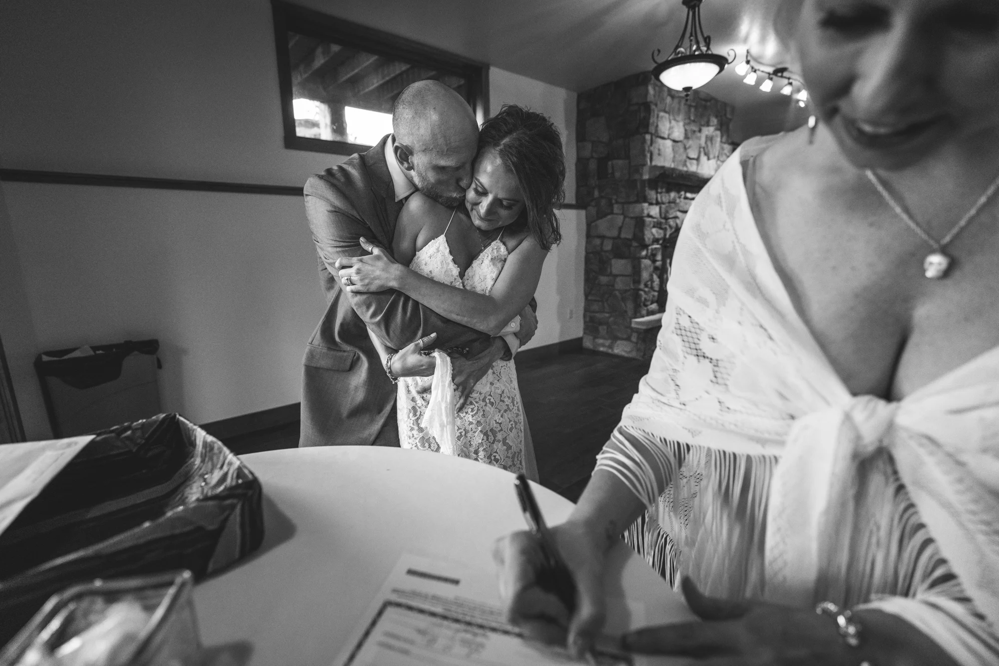 Black and white photo - a woman in the foreground signs a marriage certificate while the groom hugs his bride from the back in the background. Taken in an event room at the Black Canyon Inn in Estes Park, Colorado.
