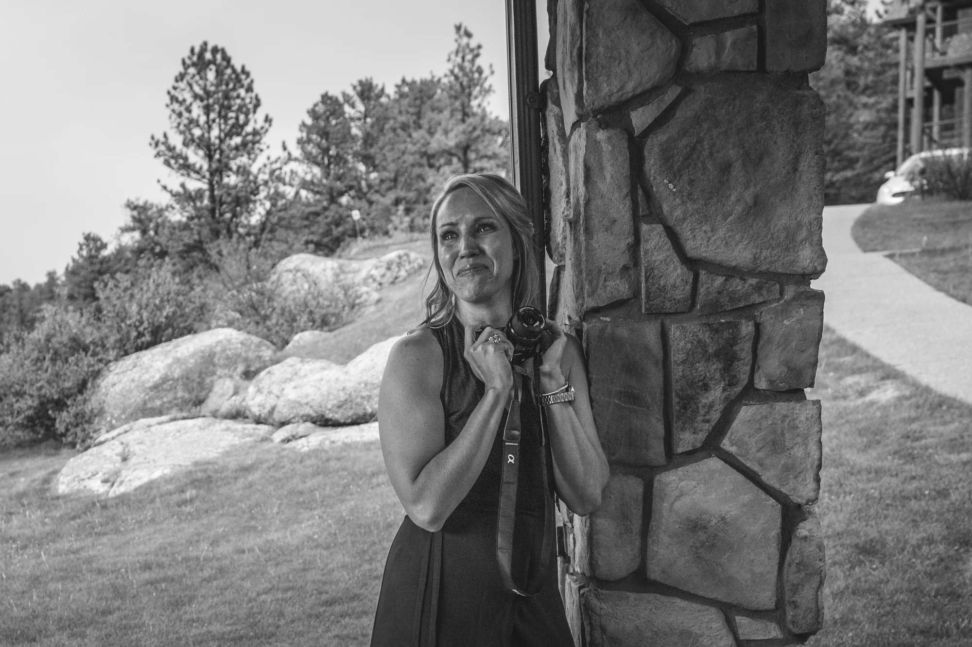 Black and white photo of a woman leaning up against a stone column with a beautiful woodsy background as she clutches a camera and cries tears of joy. Taken during a wedding ceremony in Estes Park, Colorado.