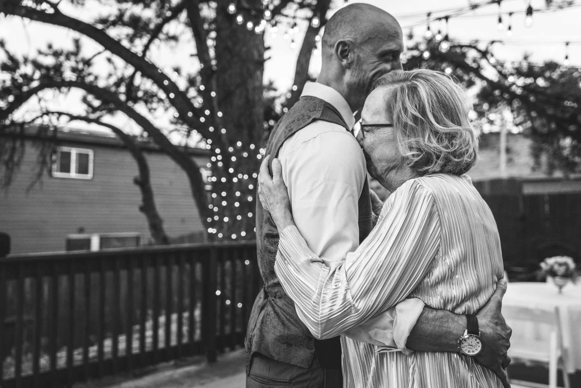 Black and white photo of an outside wedding reception with Christmas lights on a tree in the background as a mother lays her head on her son's shoulder and cries as they dance. Taken in Estes Park, Colorado.