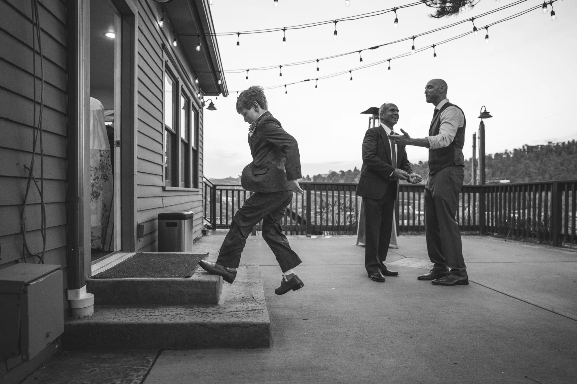 Black and white photo of a little kid at a wedding reception at the Bristlecone Inn in Estes Park, dancing on the steps outside while wearing a suit and while the groom and father-of-the-bride talk in the background.