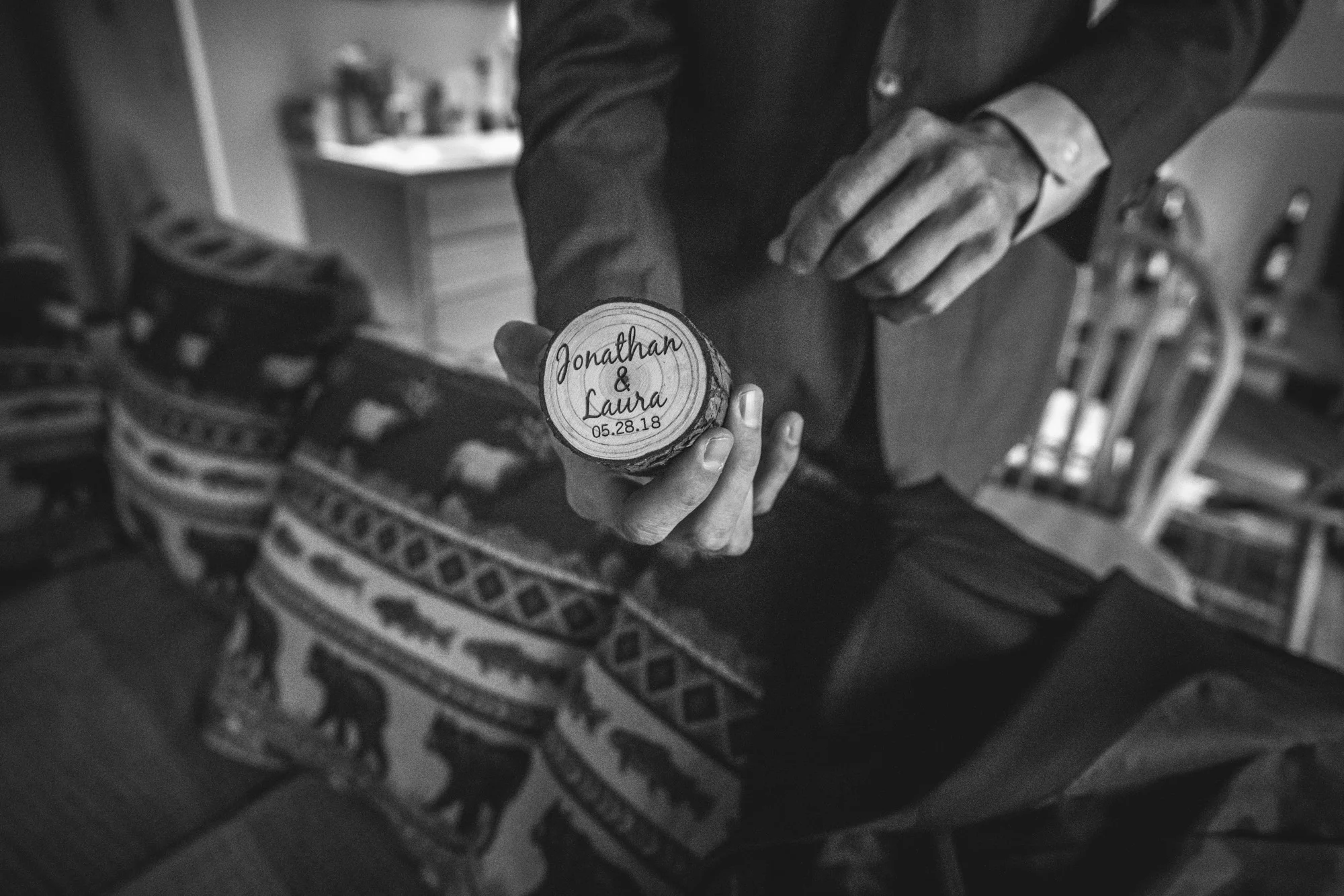 Groom holds a small, circular wooden ring box engraved with "Jonathan & Laura 05.28.18", the date of their wedding
