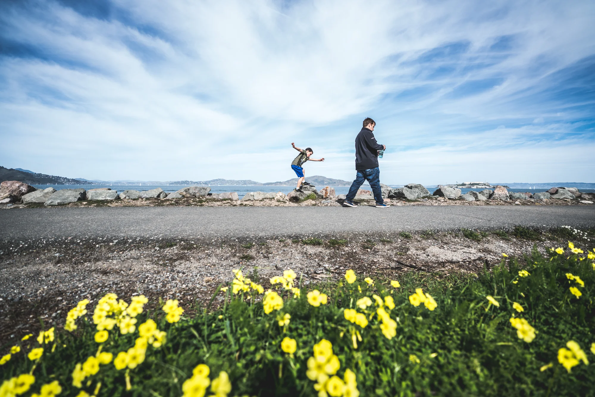 Boy jumping on rocks that line the ocean with bright yellow flowers in the foreground