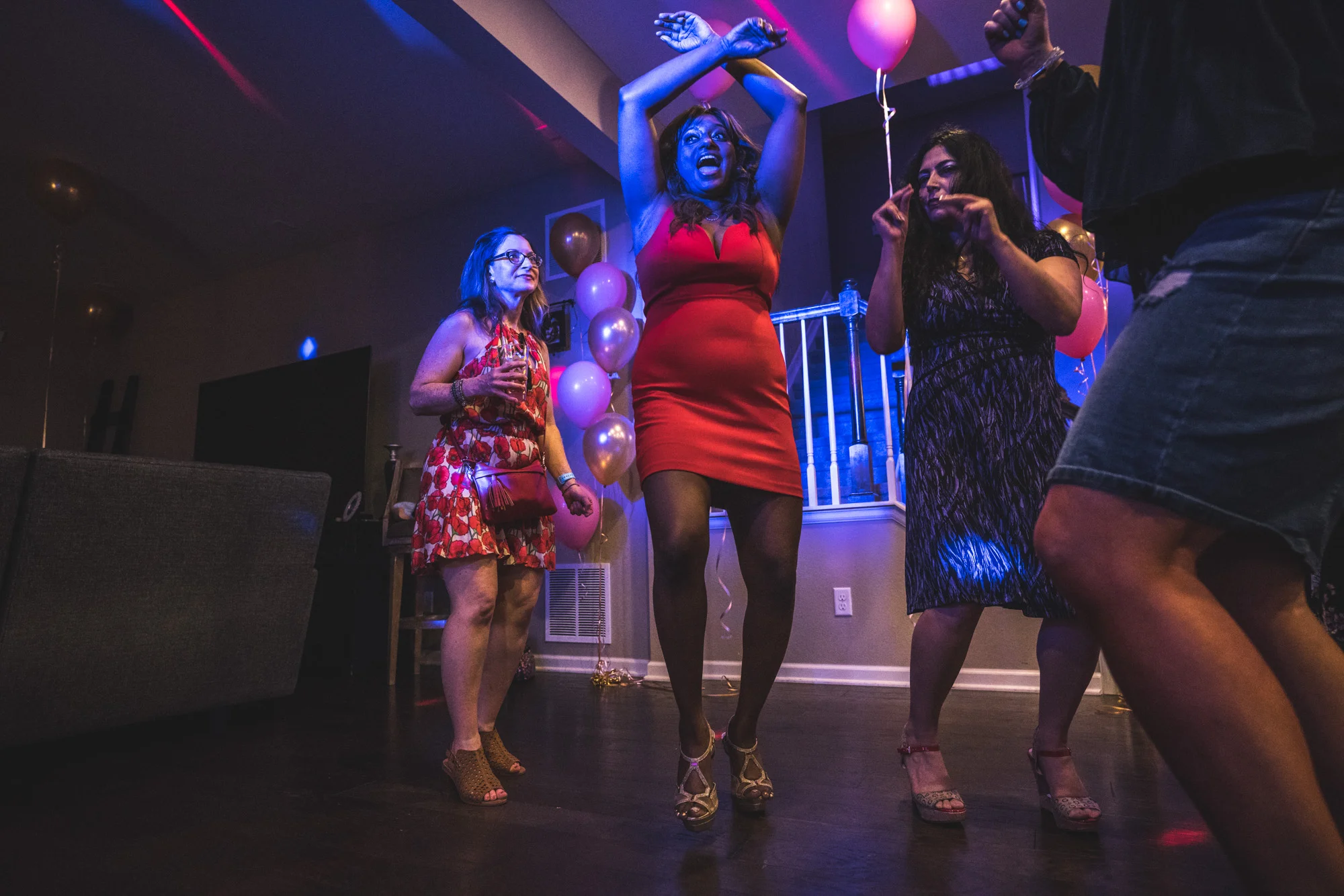 Woman in a bright red dress lifts her arms as she dances wildly at her birthday party, surrounded by bright, colorful lights and balloons