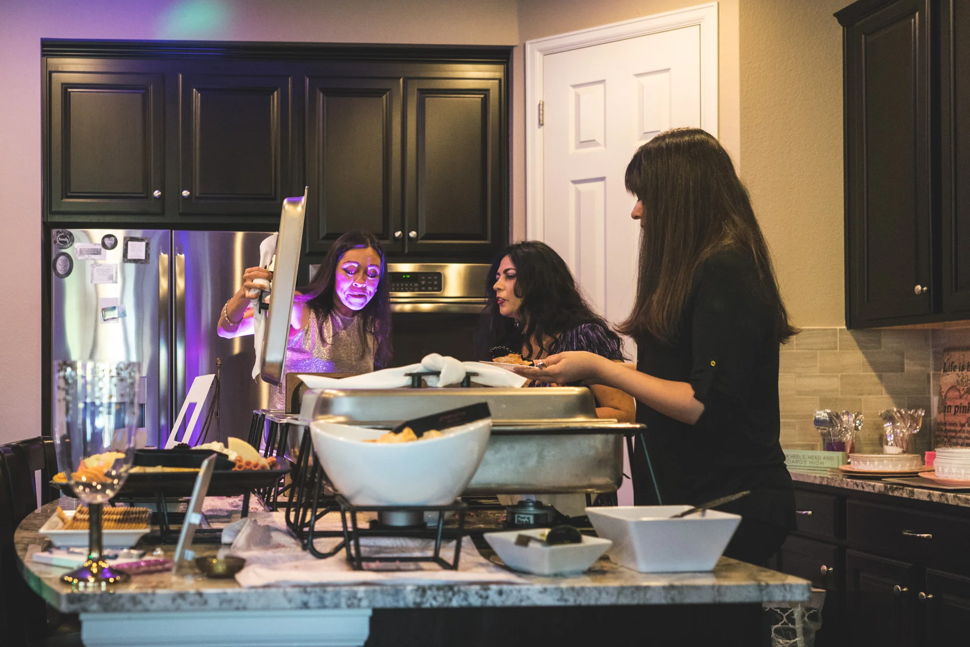 Three women look in catered dishes, one woman's face illuminated by a purple up-light.