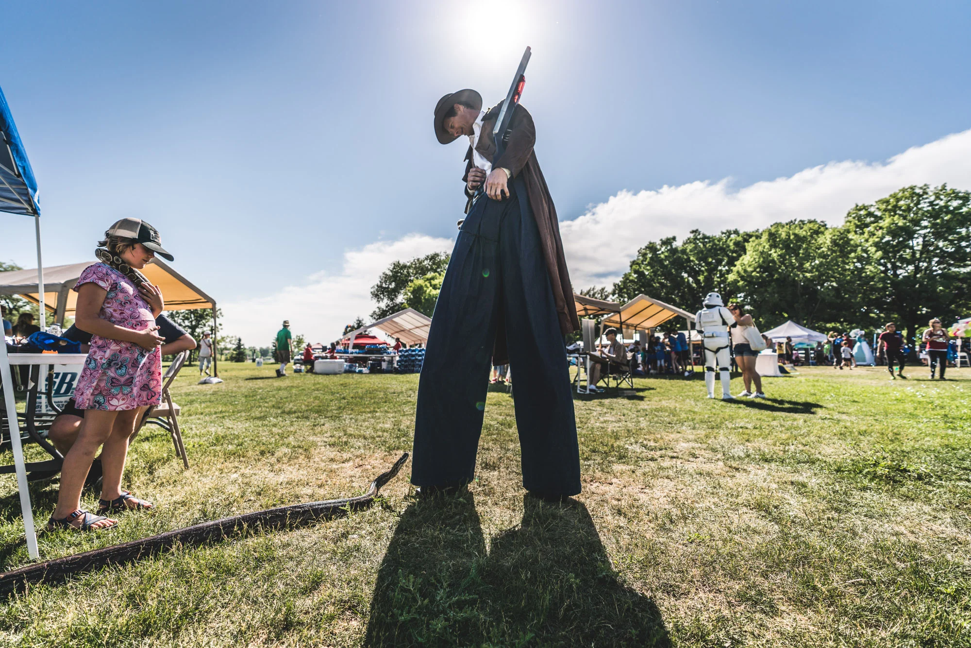 Man on stilts at an event ground looks down at a giant snake who is roaming free at the event